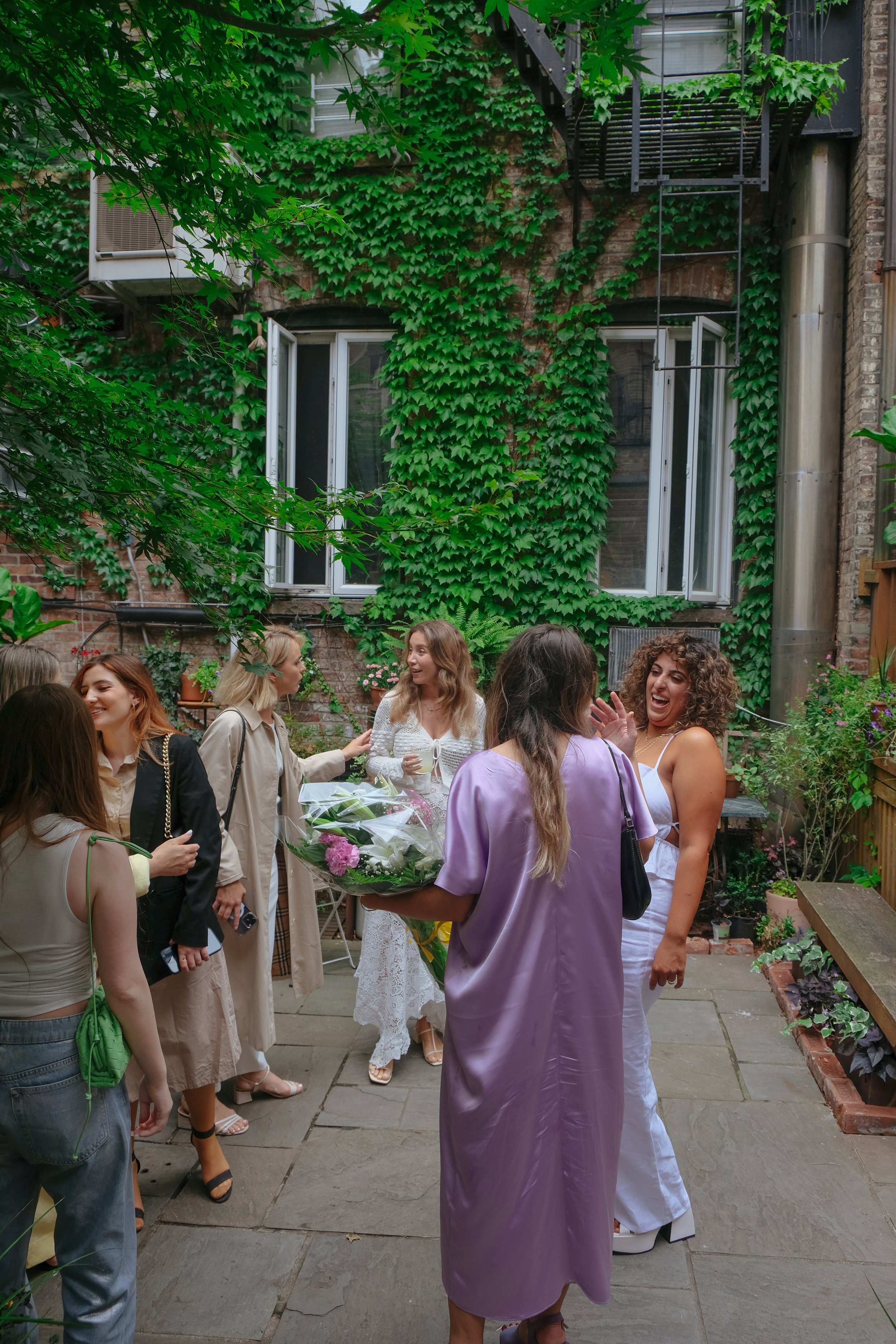 Group of women socializing in a lush green backyard with ivy-covered brick wall, open windows, and outdoor decor.