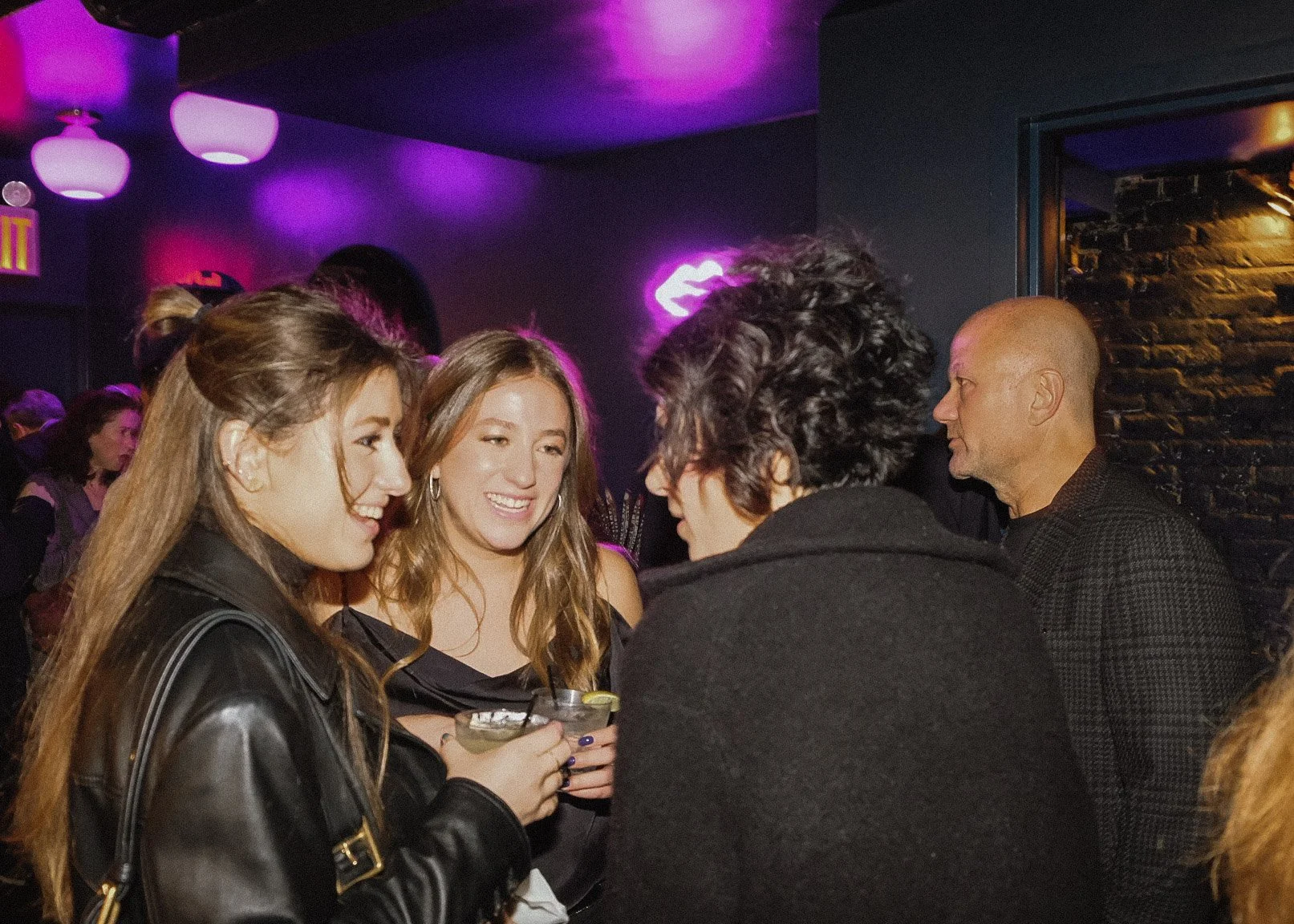 Group of four people talking and smiling at a dimly lit bar or club with purple neon lights.