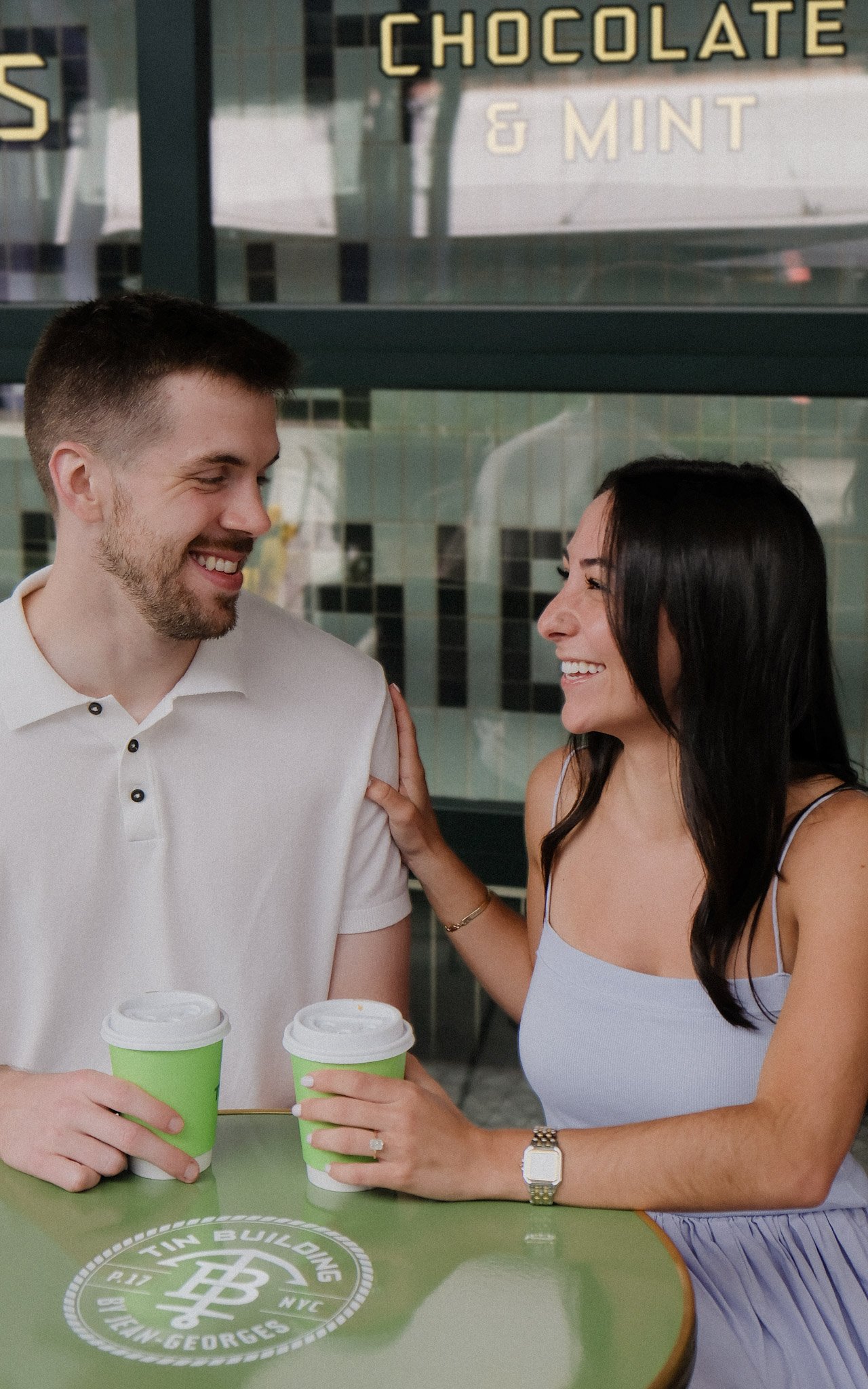 A smiling couple with drinks in green cups sitting at a green table outside a cafe with a sign that reads 'Chocolate & Mint'.