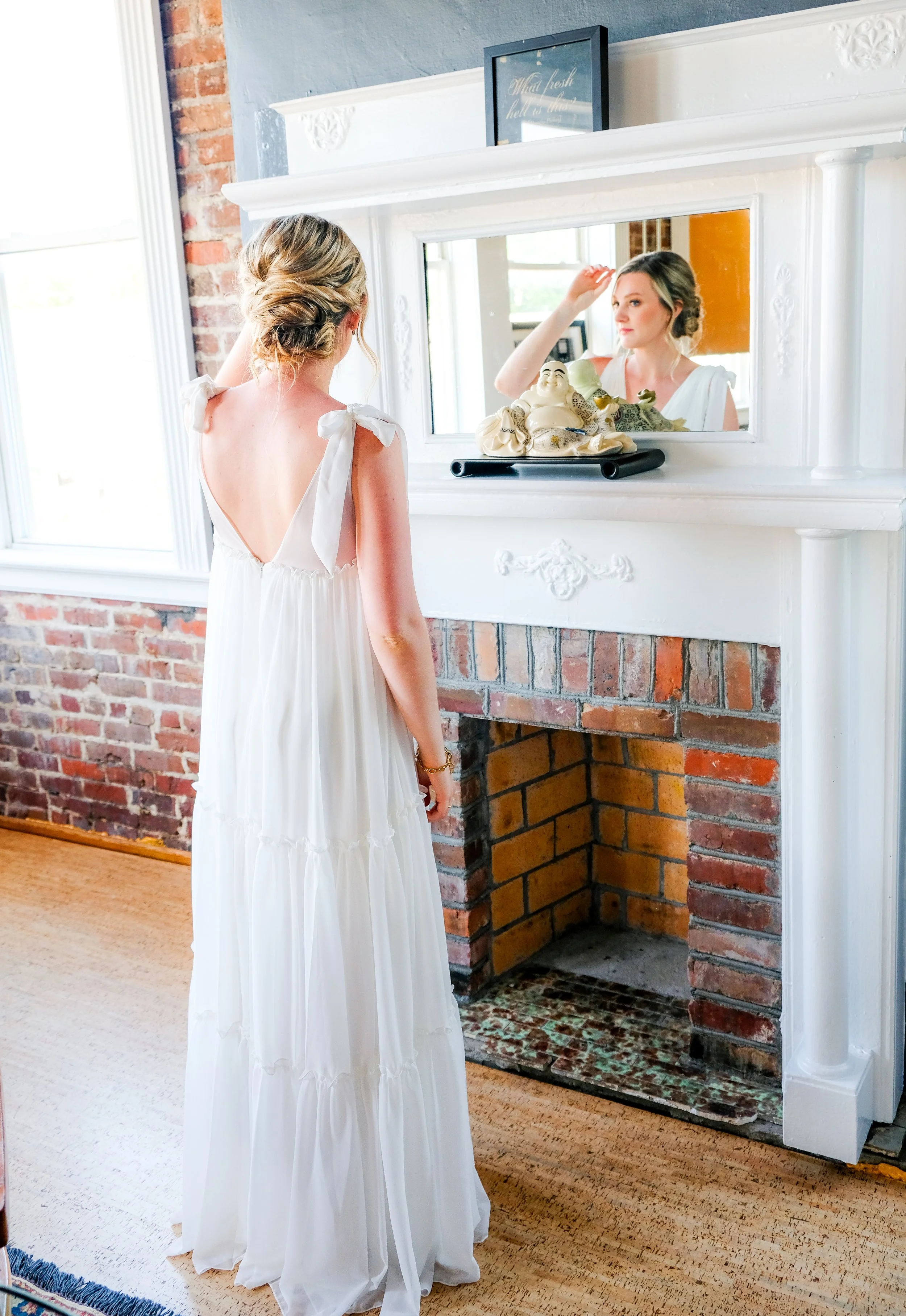 A woman in a white dress with tied shoulder straps is looking at her reflection in a mirror above a fireplace. The fireplace is made of brick, with a white mantel. The room has a large window with white trim, and there is a framed quote above the mir