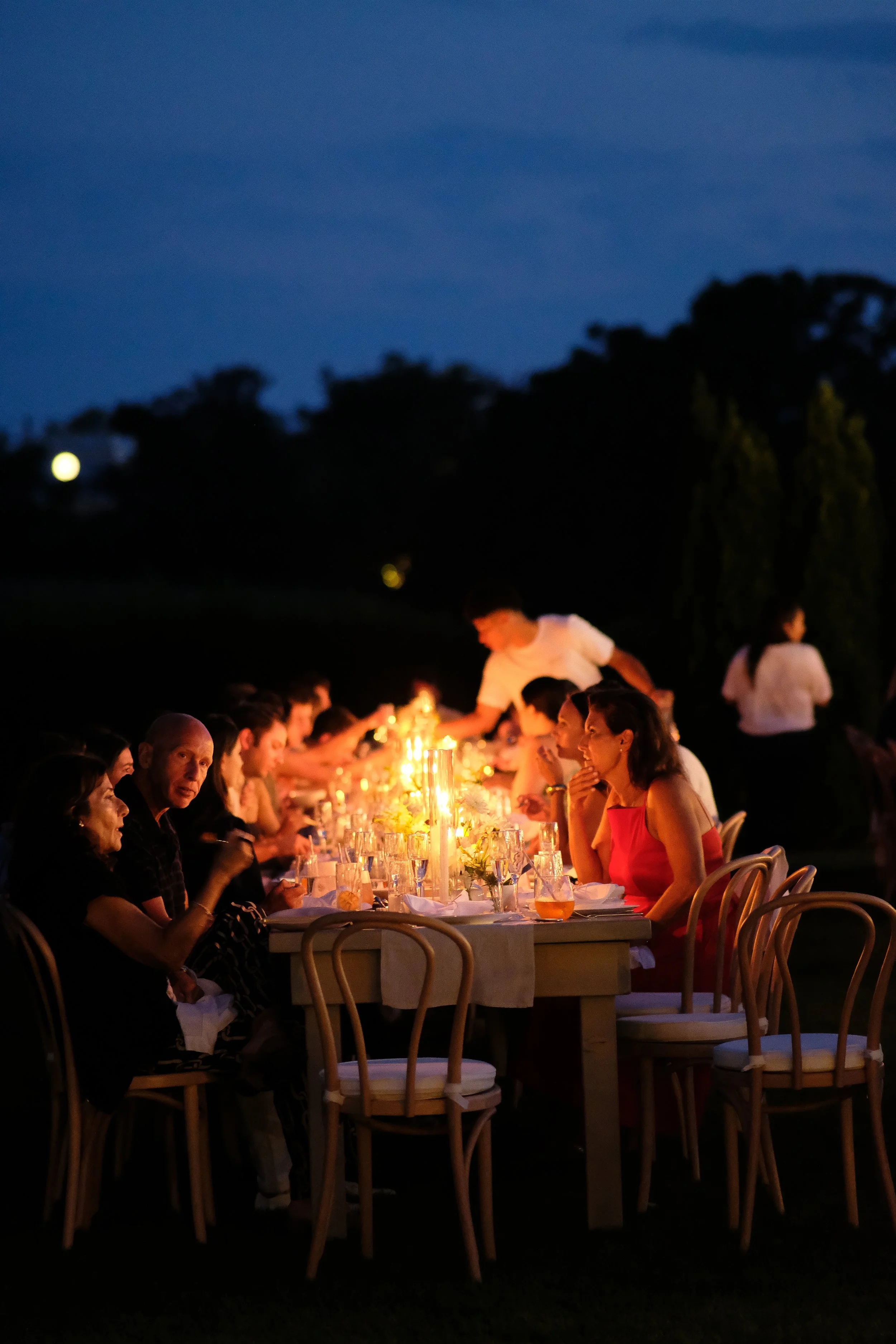People dining at an outdoor candlelit dinner table during evening with a dark sky in the background.