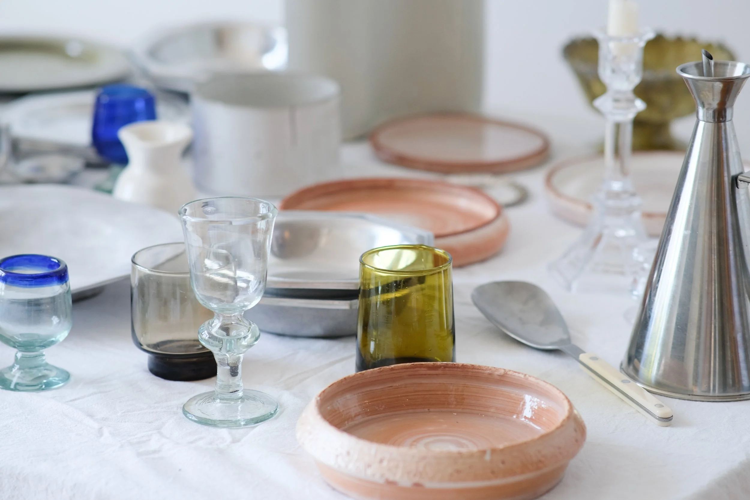 Various glassware, plates, bowls, and utensils arranged on a white tablecloth, including a green drinking glass, clear wine glasses, a spatula, and a metal pitcher.