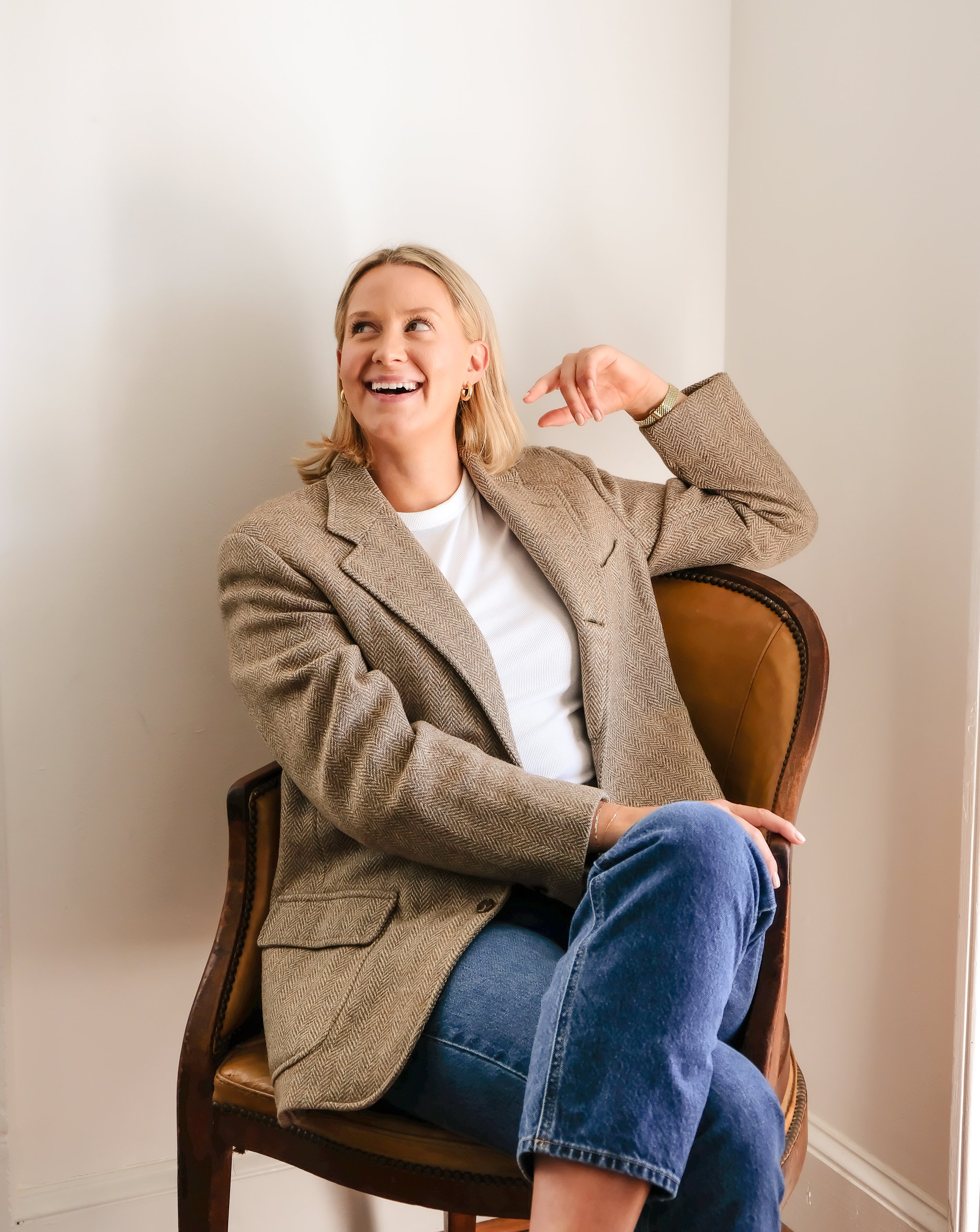 A woman with blonde hair sitting on a brown leather chair, smiling and gesturing with her fingers near her ear, wearing a beige blazer, white top, and blue jeans, in a plain room.