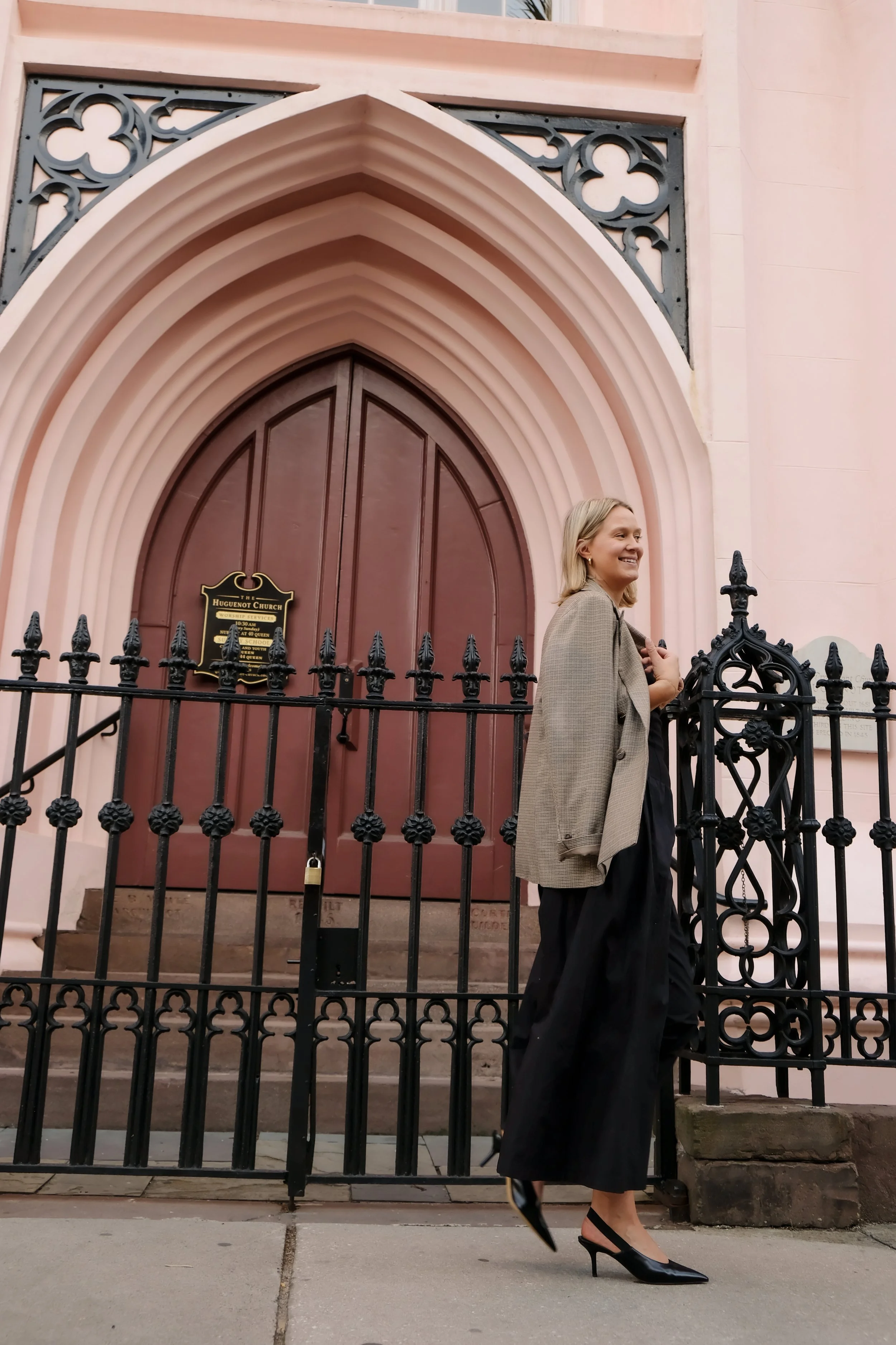 A woman in black high heels, black wide-leg pants, a gray blazer, and a dark top standing outside a pink church with arched doorway and black decorative iron fence.