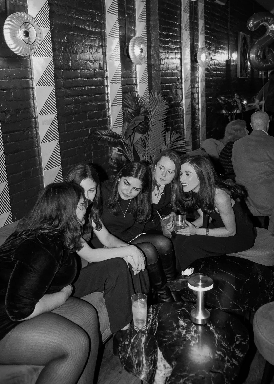 A group of four women sitting on a sofa at a bar or lounge, engaged in conversation, with drinks in their hands. The background has a brick wall, decorative lights, plants, and other patrons.