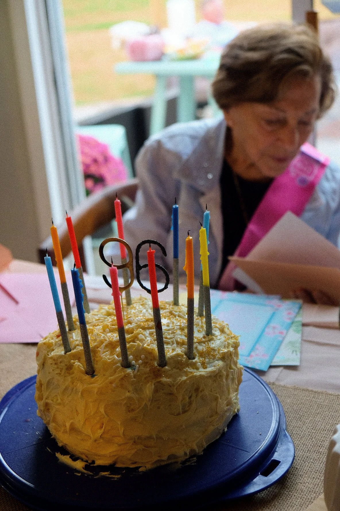 A birthday cake with lit candles and a number '36' topper on a table, with a woman in the background reading a card.
