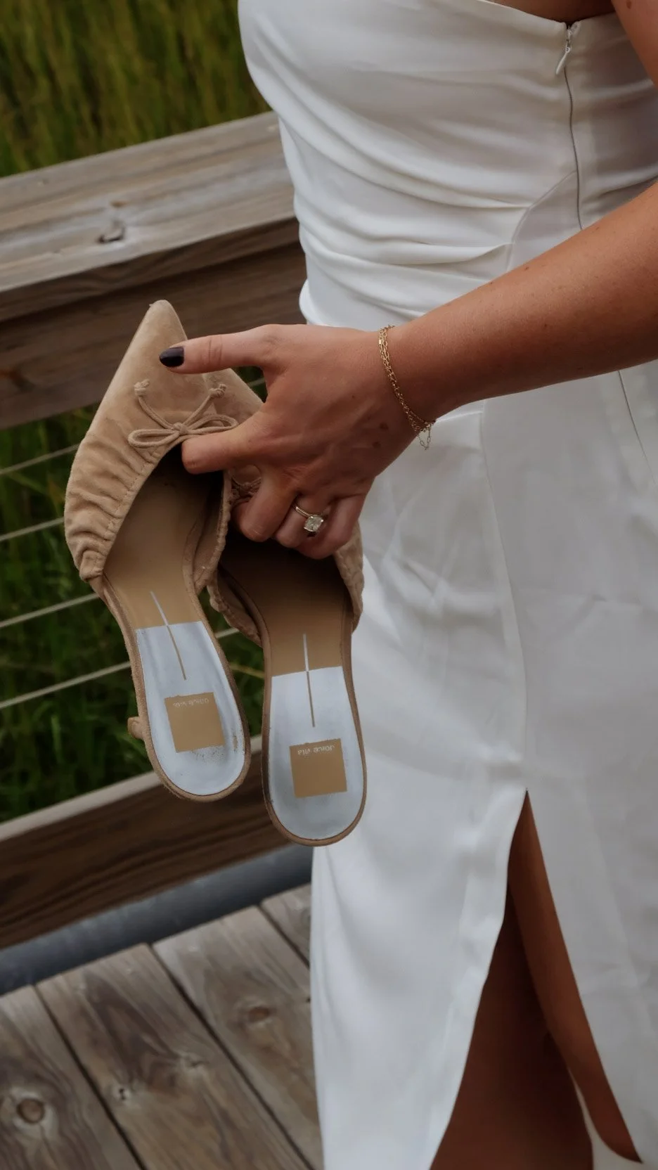A woman in a white dress holding a pair of beige ballet flats, standing outdoors on a wooden deck with greenery in the background.