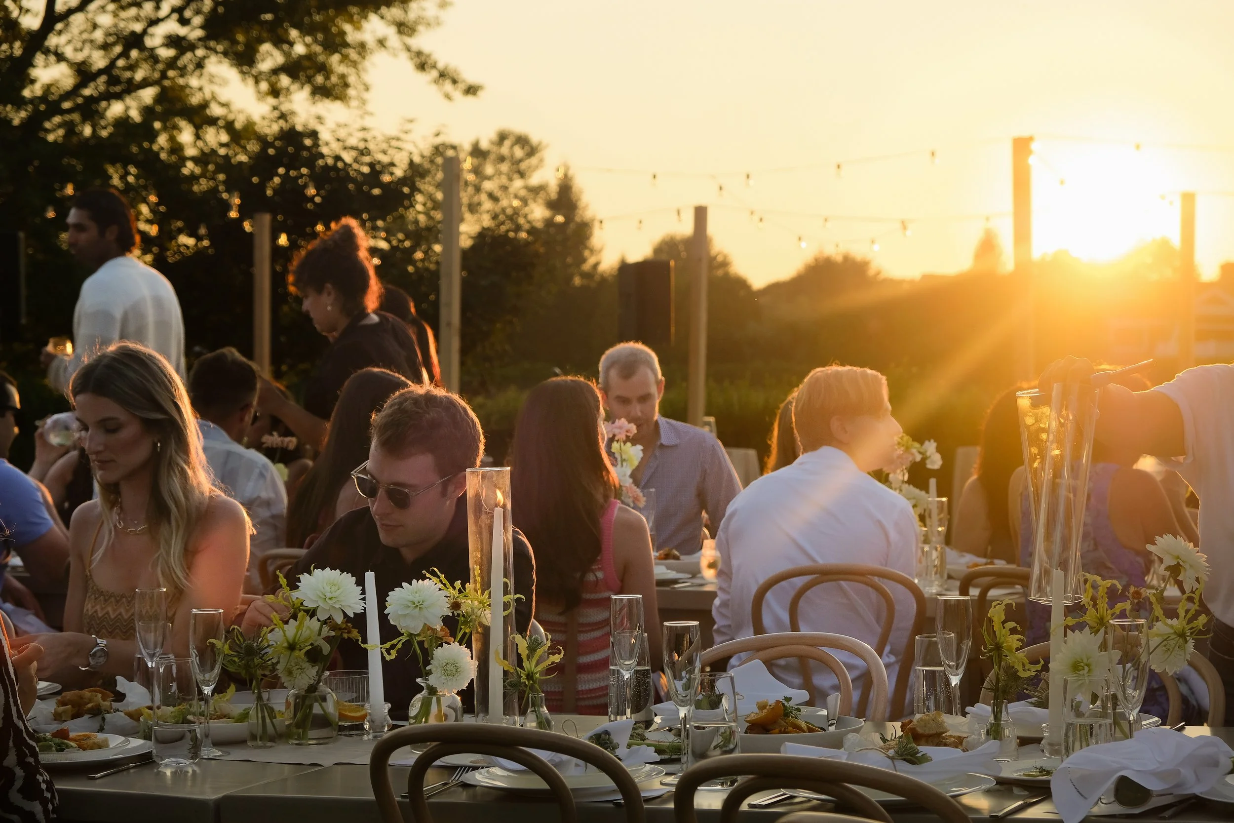 People gathered at an outdoor evening event, sitting at decorated tables with flowers, enjoying food and drinks, with the sunset in the background.