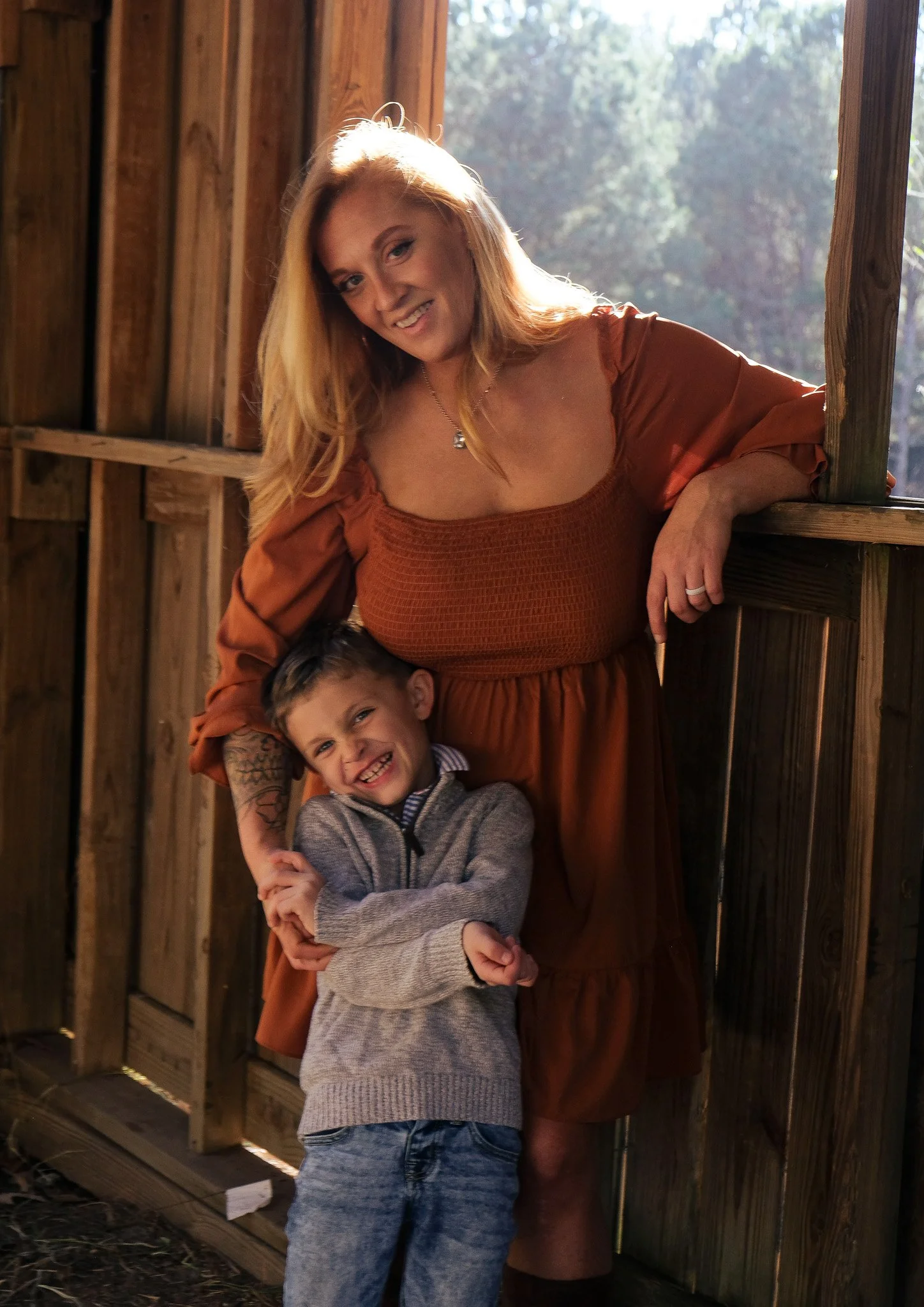 A woman with long blonde hair and a young boy with short dark hair, smiling and embracing each other in a wooden outdoor structure, sunlight shining through the trees.