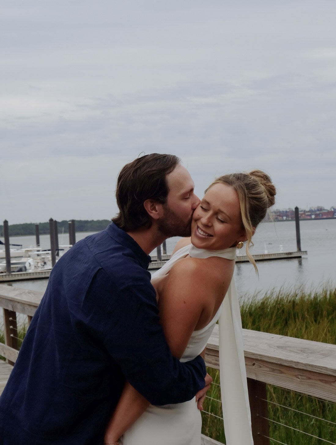 A couple embracing and sharing a kiss near a body of water, with a dock and boats in the background.