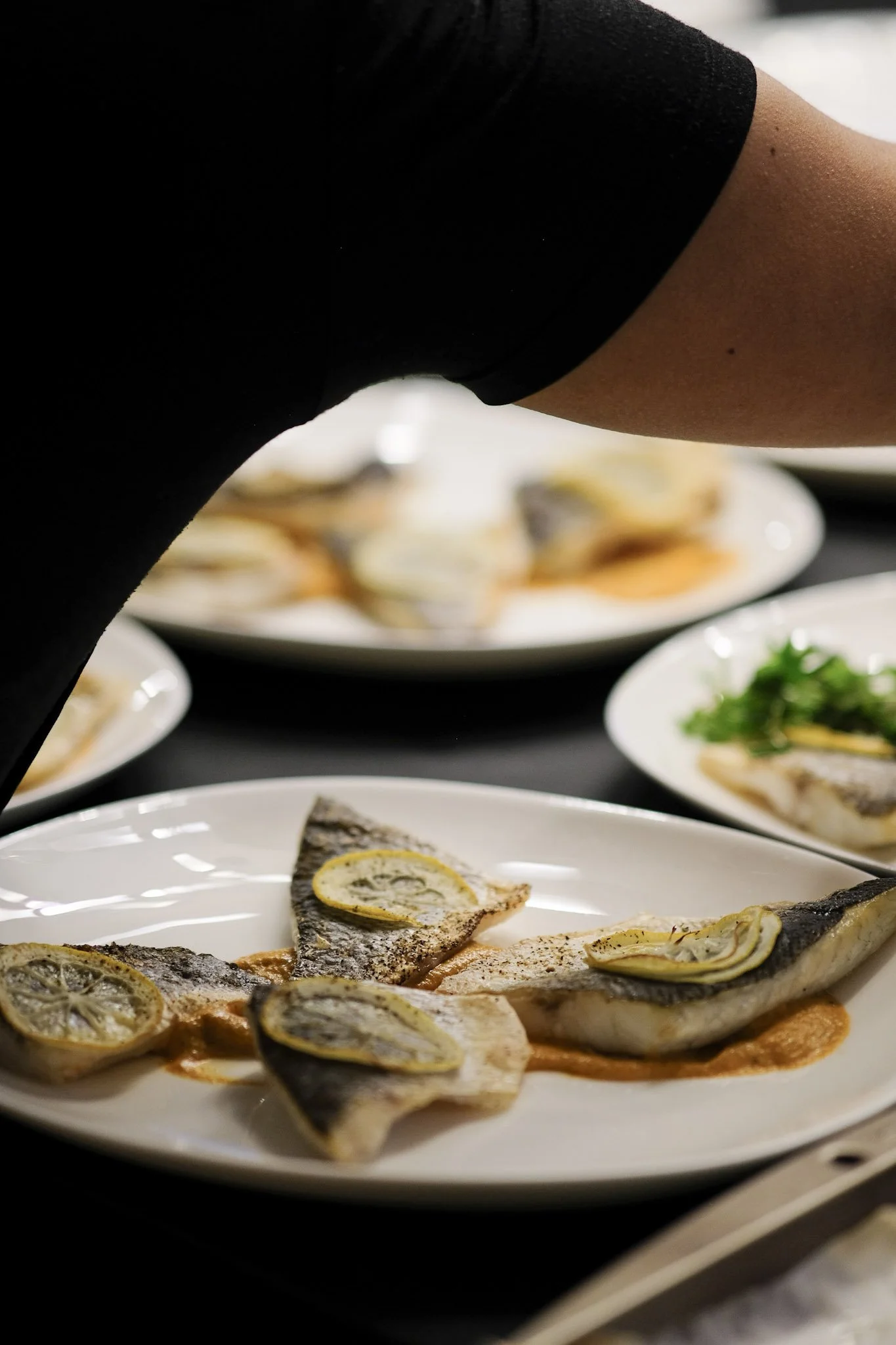 Close-up of a person preparing grilled fish garnished with lemon slices on a white plate, with additional plates of food in the background.