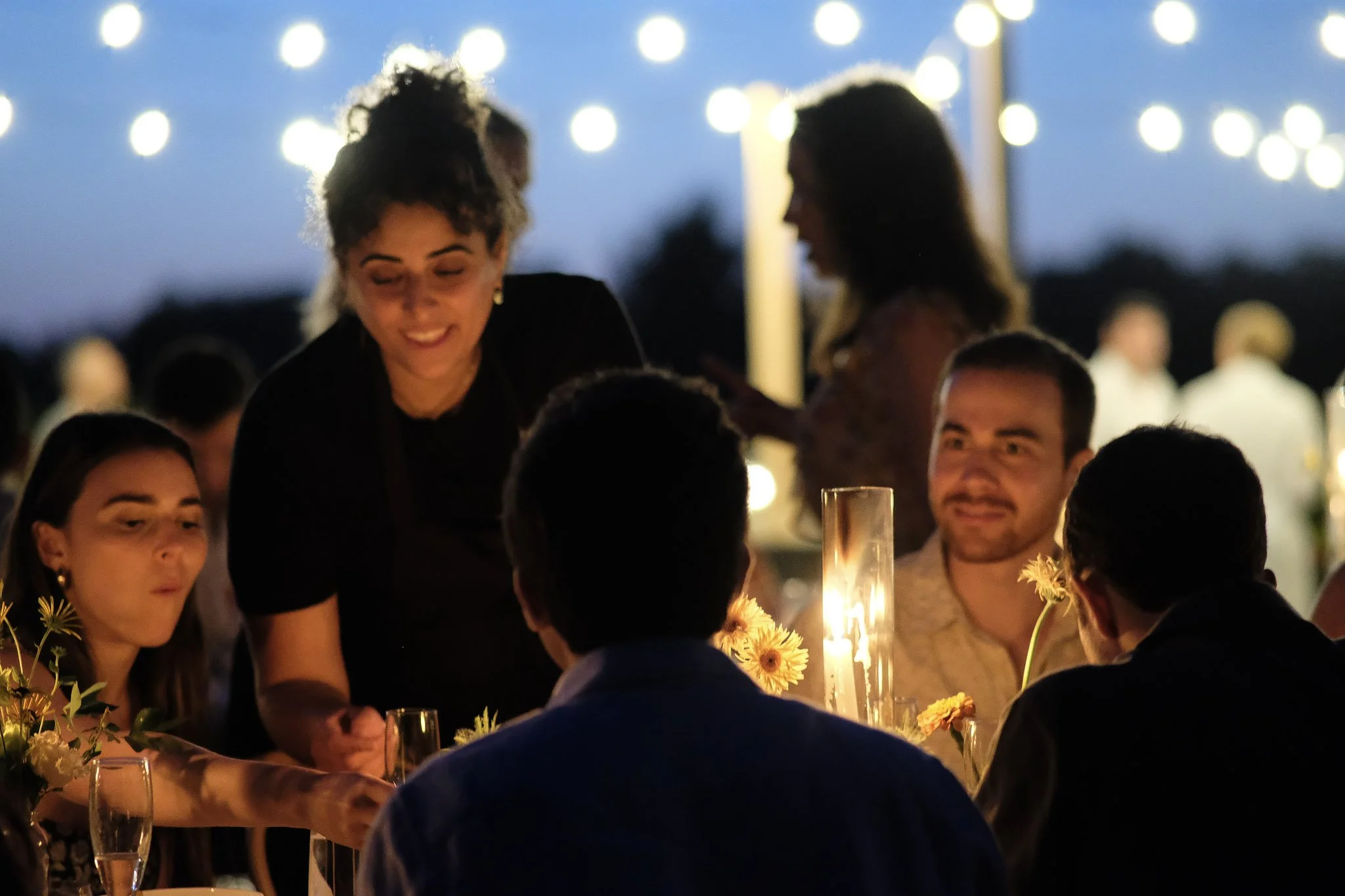People sitting around a table with candles and flowers, enjoying an outdoor dinner in the evening, with string lights overhead.