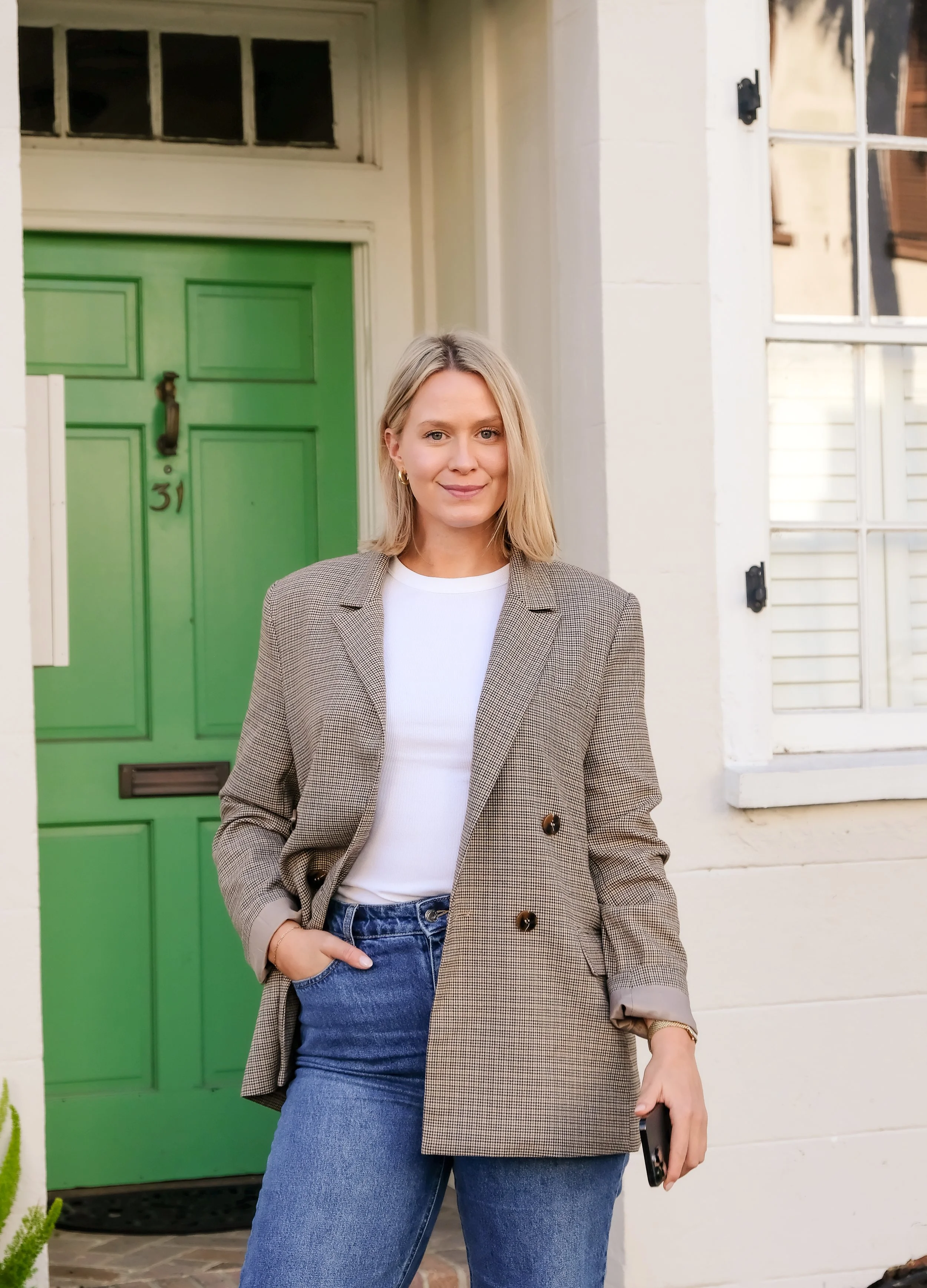A woman standing outside a house with a green door, wearing a beige blazer, white t-shirt, and blue jeans, holding a phone in her right hand, smiling at the camera.