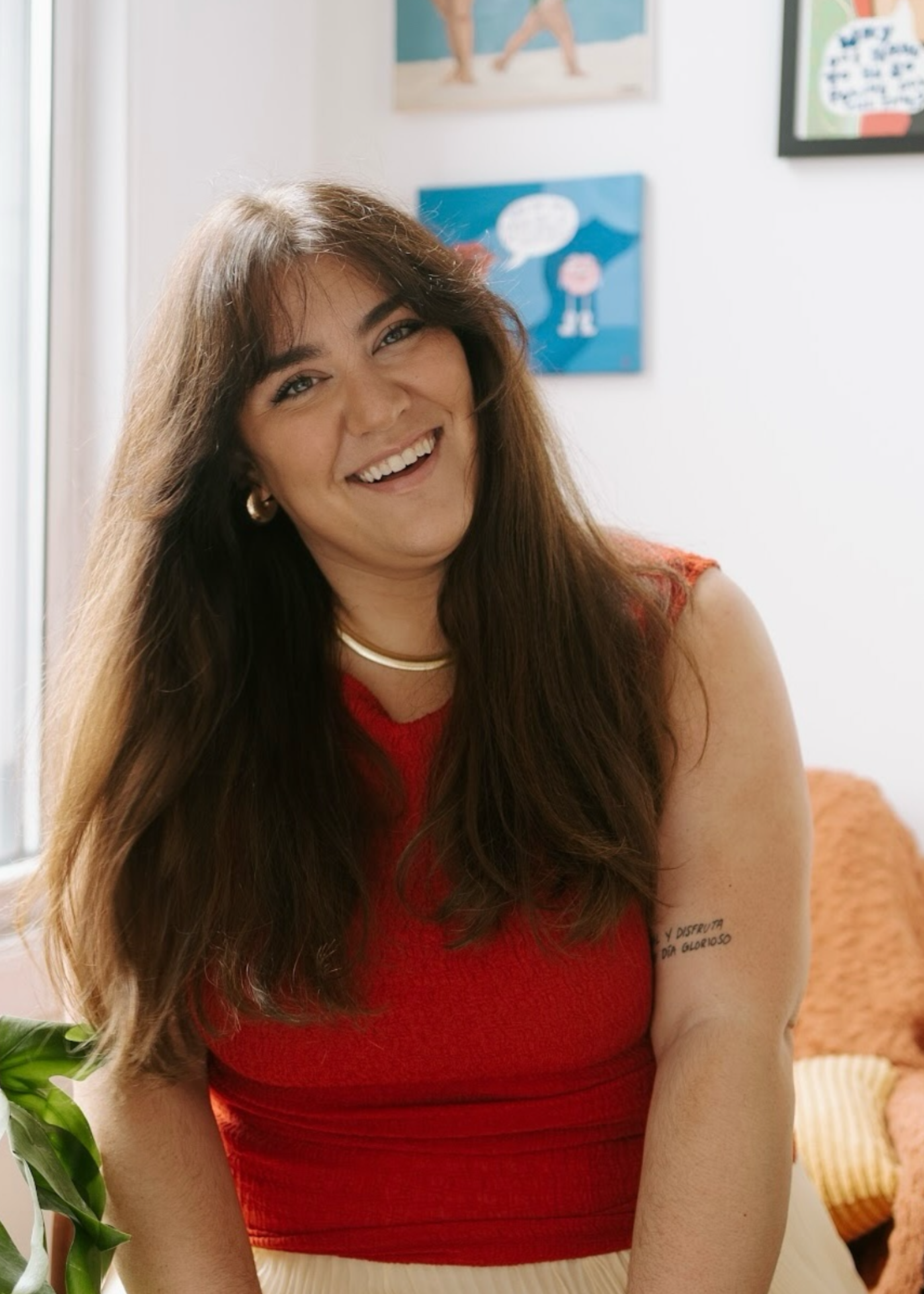 A woman with long brown hair, wearing a red sleeveless top, smiling and looking at the camera, seated inside a room with colorful children's artwork on the wall behind her.