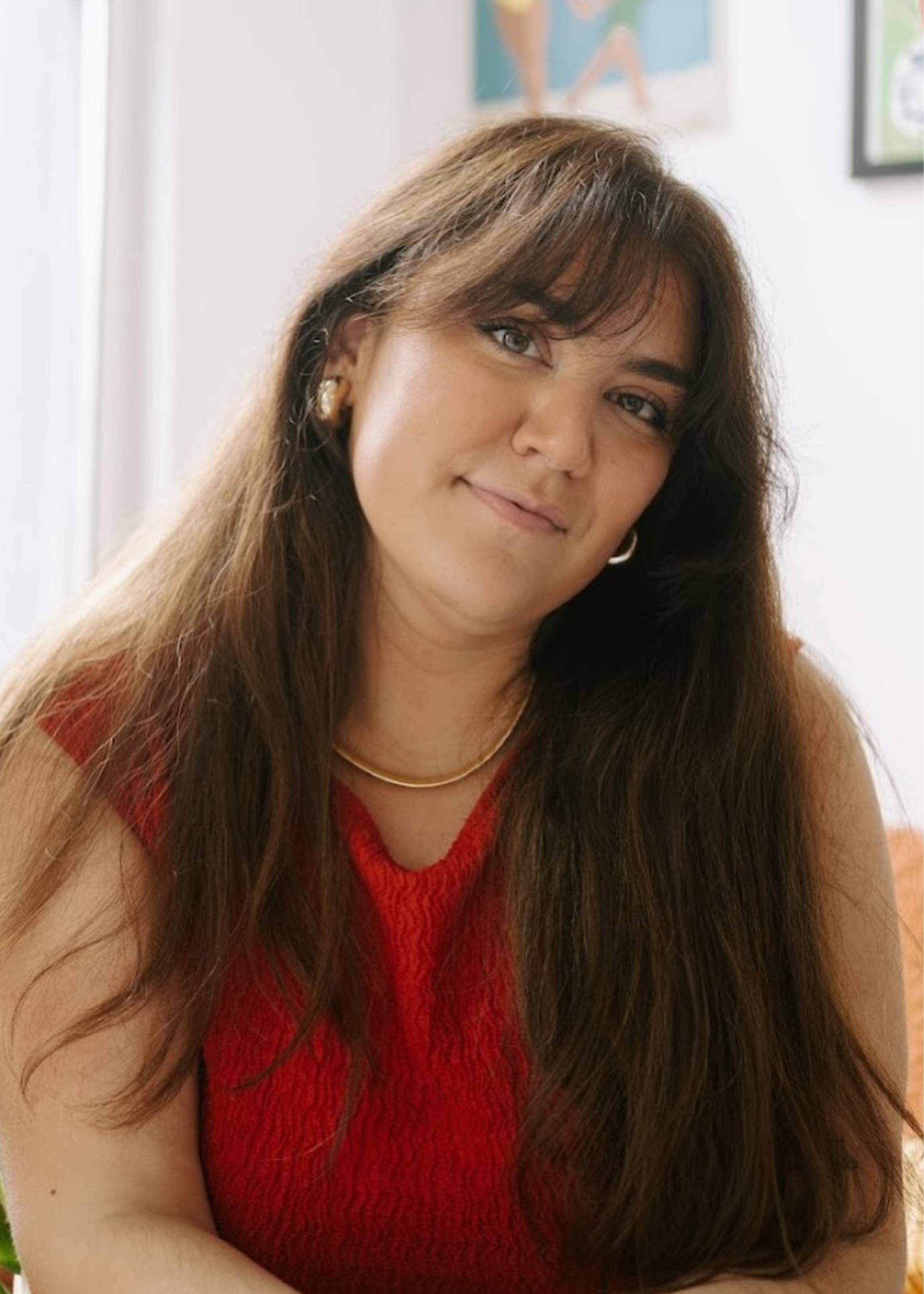 A woman with long brown hair, wearing a red sleeveless top and gold jewelry, sitting indoors with a colorful abstract painting and a window in the background.