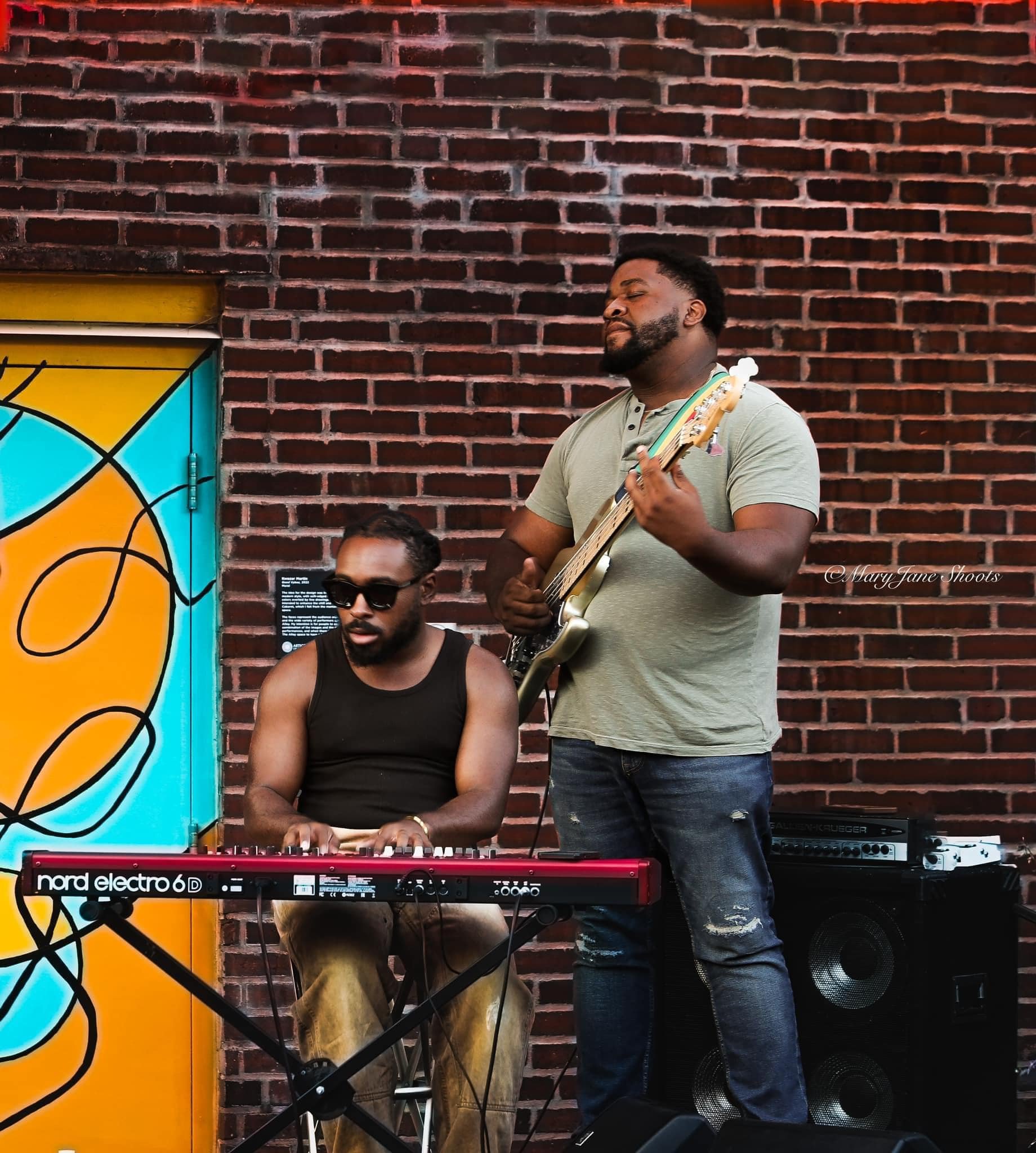 Two male musicians performing outdoors against a brick wall. One playing a keyboard and the other with an electric guitar, with a colorful painted background nearby.