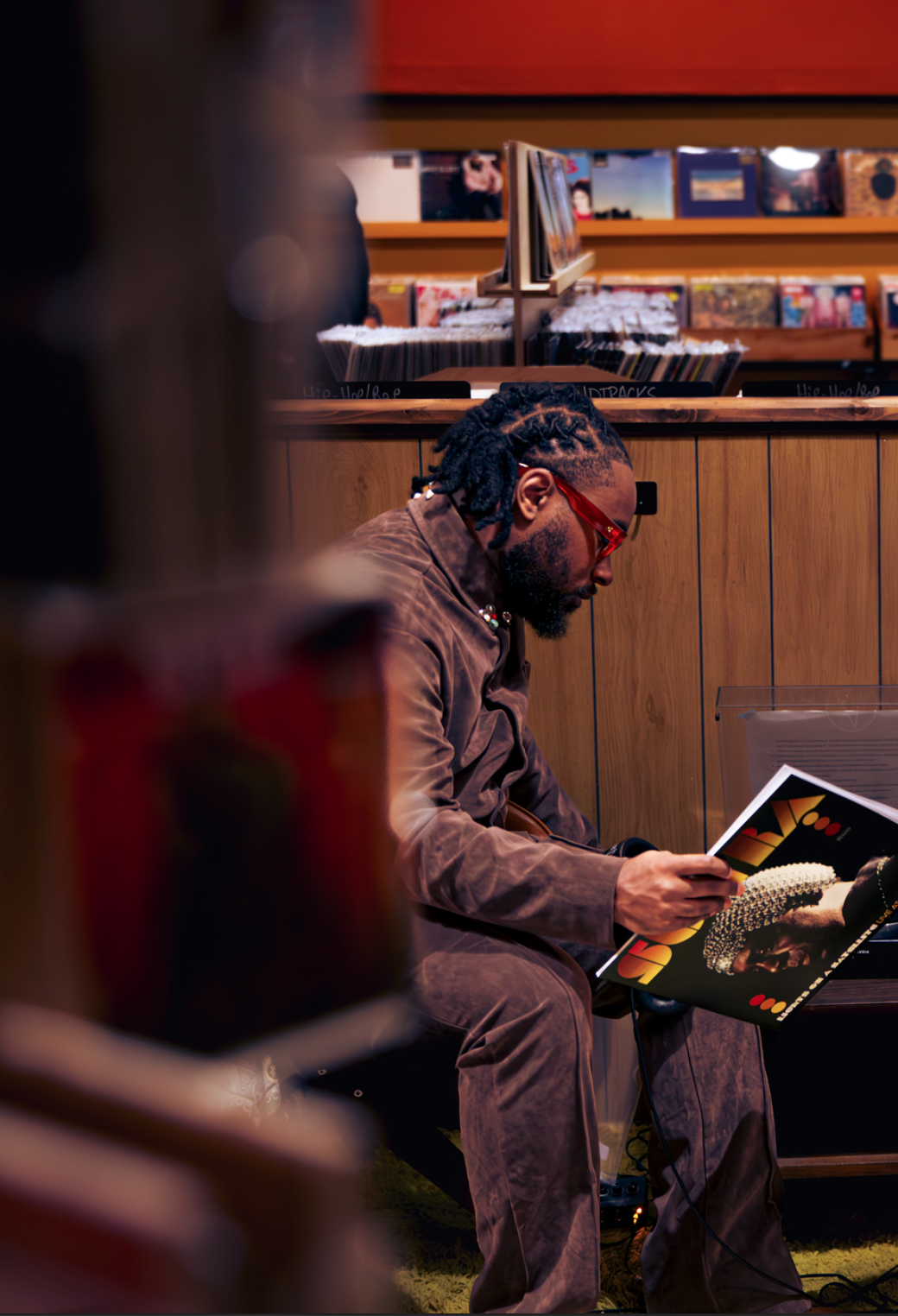 A man with dreadlocks and red glasses sits in a record store, reading a vinyl record cover.