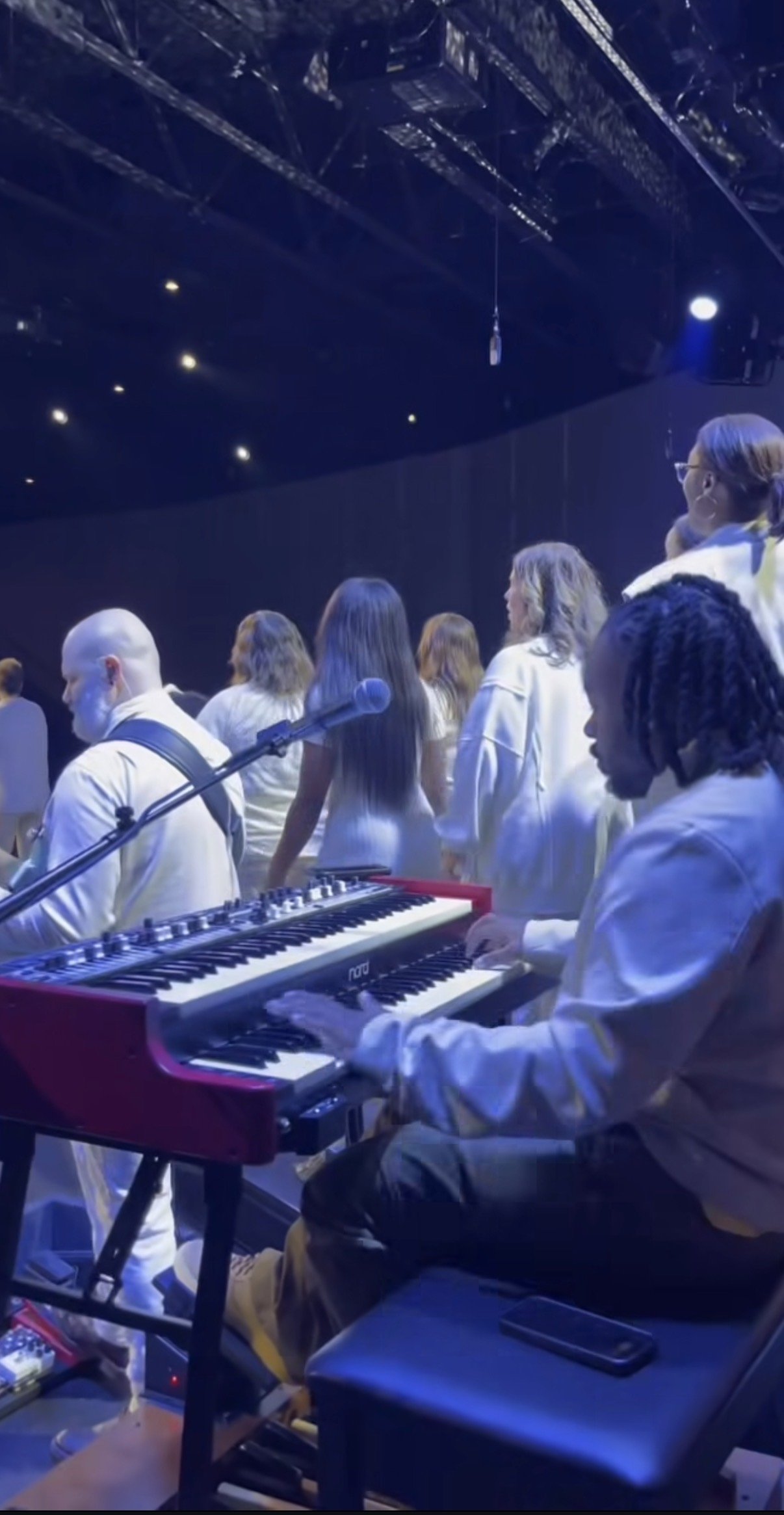 Musicians and singers on stage during a performance, with a keyboard player in focus and vocalists facing the audience, illuminated by stage lighting.