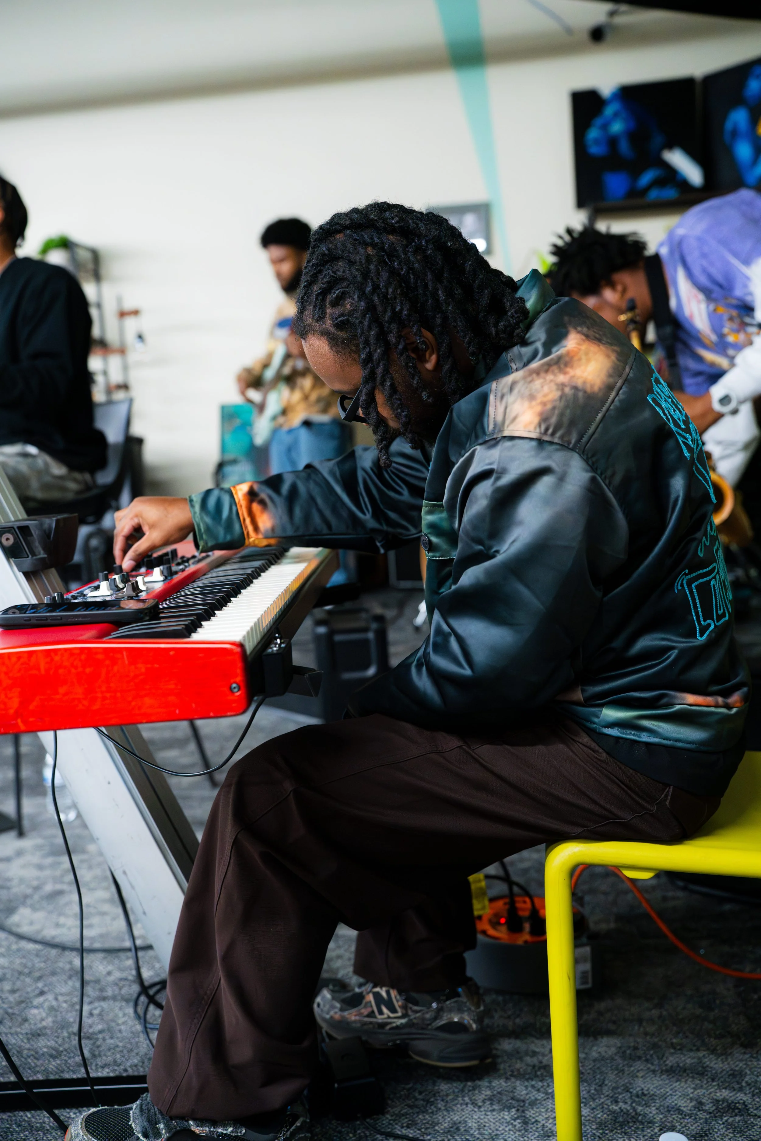 A musician with dreadlocks and glasses playing a red keyboard on a stand during a live performance, with other band members in the background.