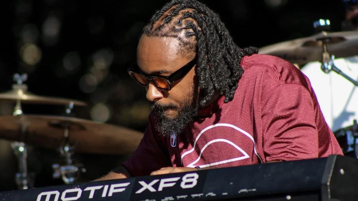 A man with dreadlocks and sunglasses playing a keyboard during a live performance.