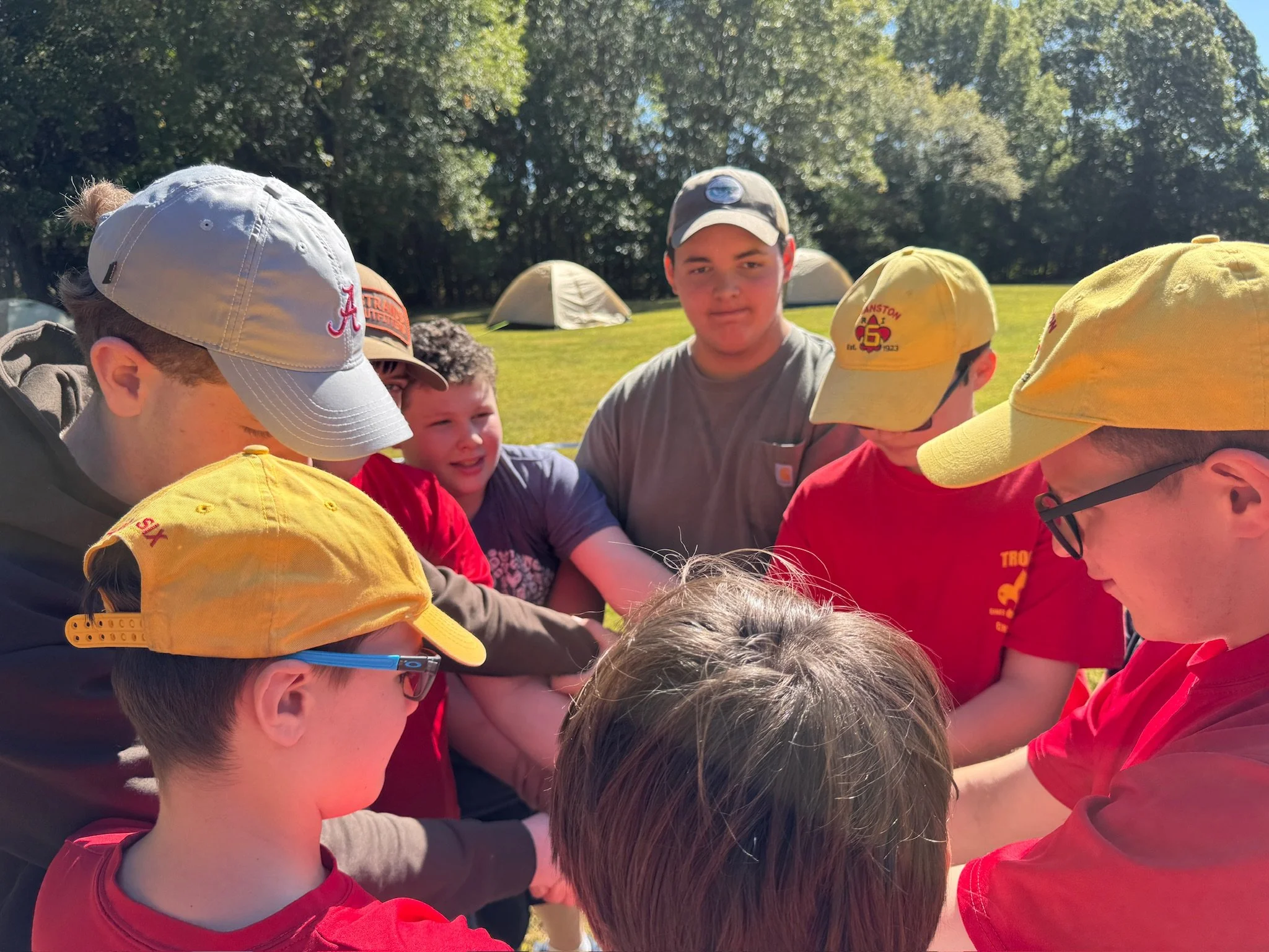 A group of young boys and teenagers huddle together outdoors, with tents and trees in the background during fall leadership training for our Scout Led program