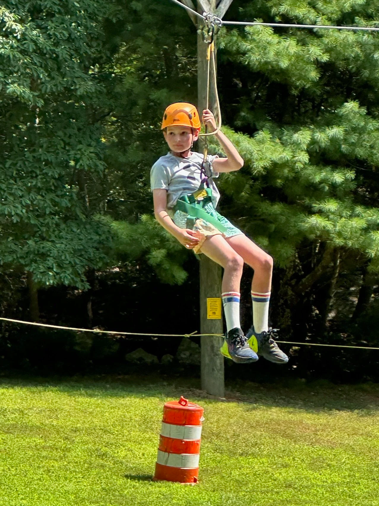 A young boy in a helmet is riding a zip line above the ground, wearing a harness, with a bright orange safety helmet, grey T-shirt, shorts, and sneakers, as he approaches a bright orange and white traffic barrel.