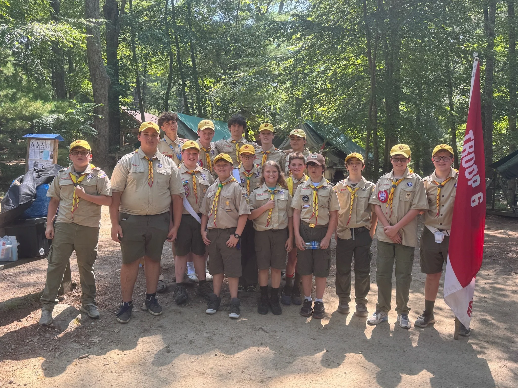 Boy and girl scouts pose for a group photo in a wooded outdoor setting, some in uniform, with a red and white flag and tents in the background.
