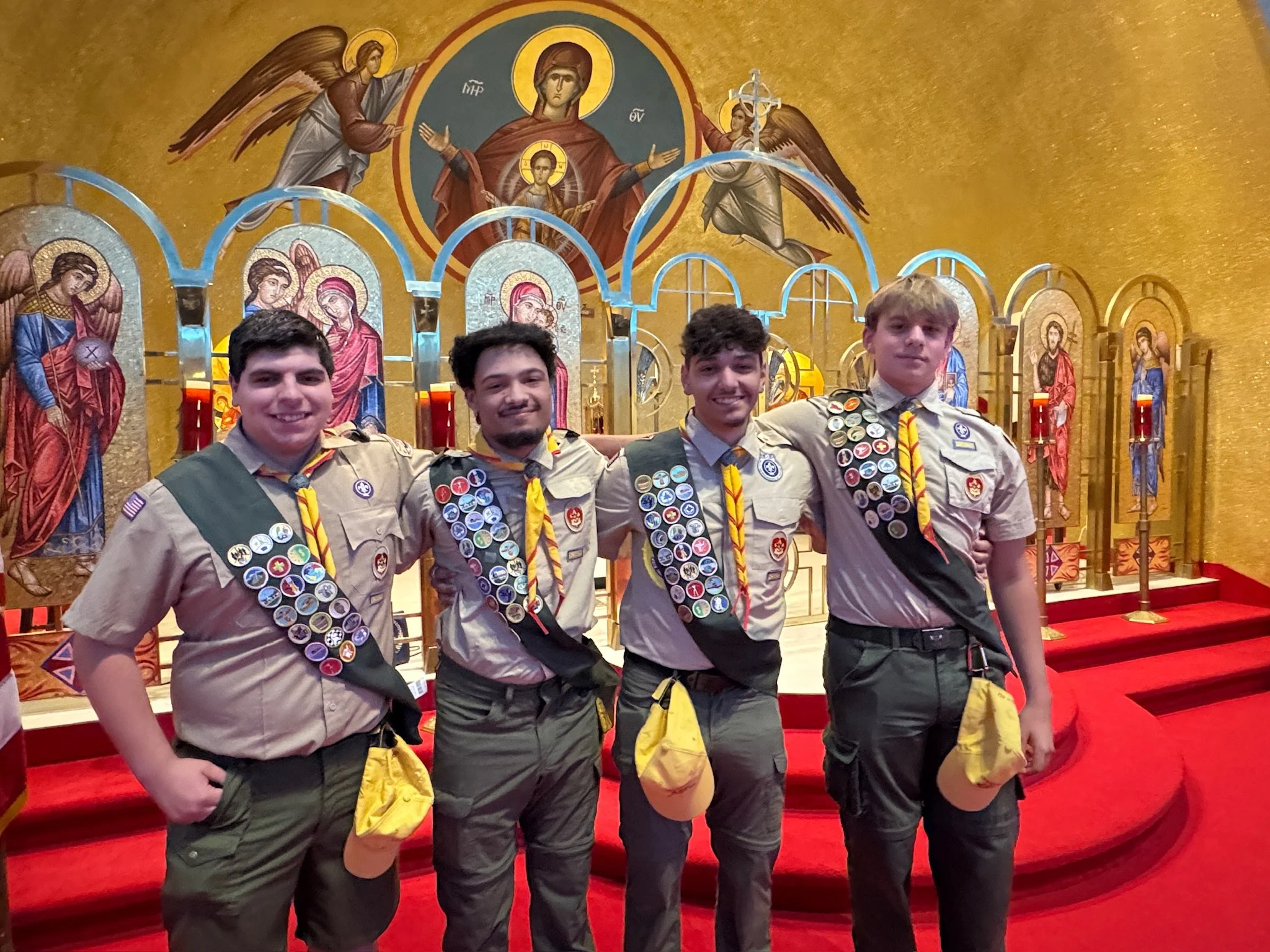 Four Boy Scouts in uniform standing in front of religious icons and a gold-painted backdrop with an image of Jesus Christ with outstretched arms and angels, inside a church or chapel.