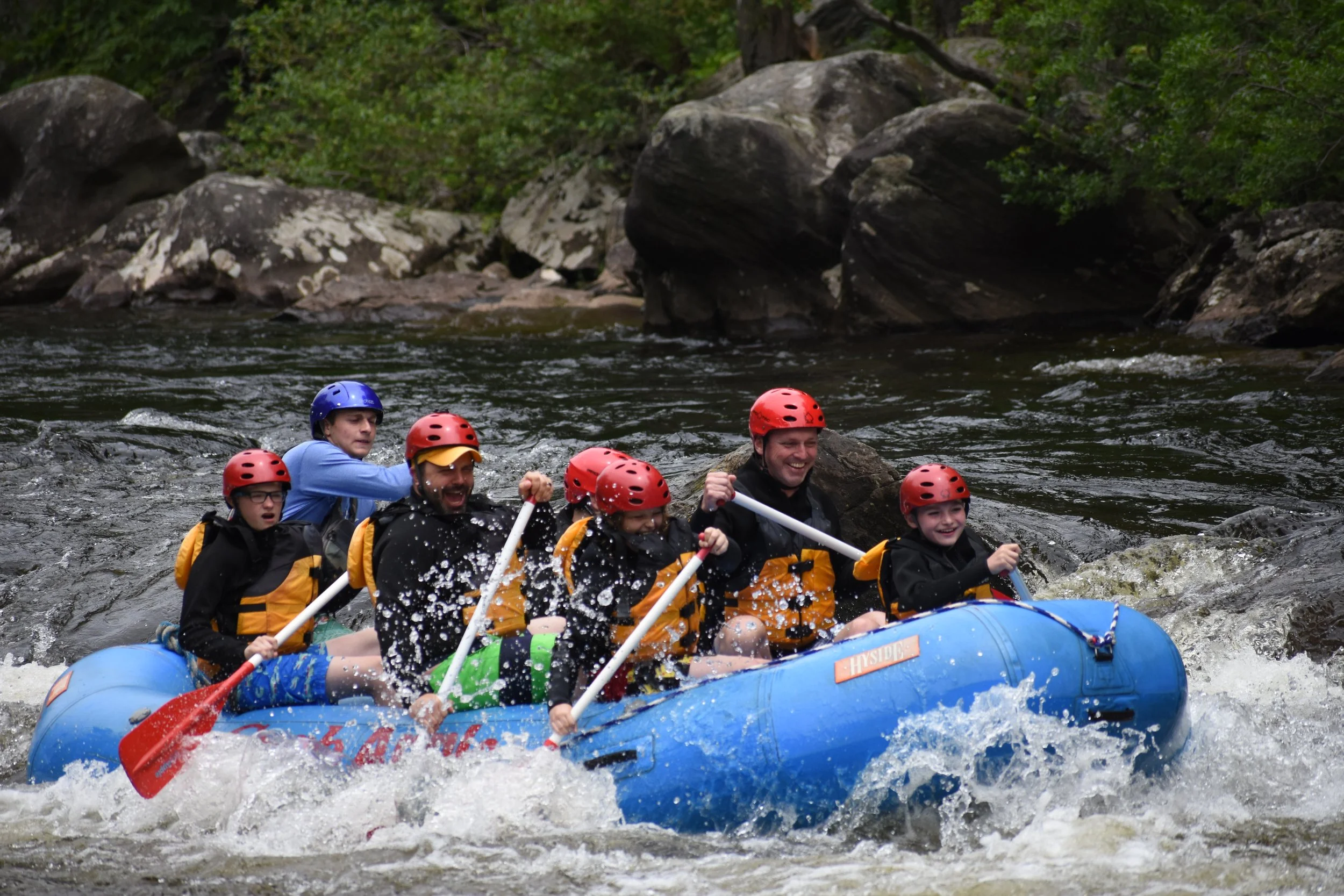 Group of Scouts and leaders white water rafting on river, wearing helmets and life jackets, with smiles and excited expressions.