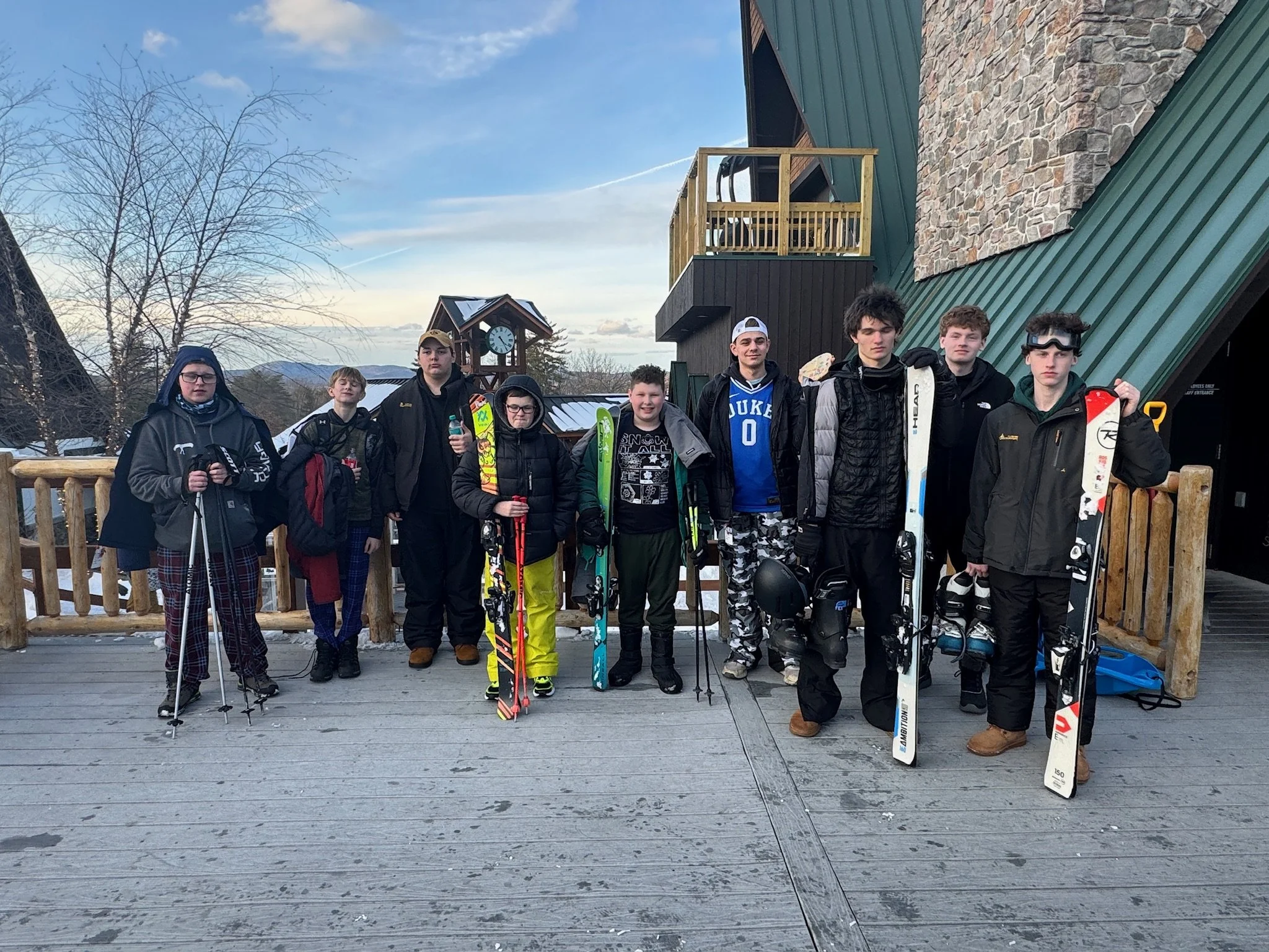 Group of young people in winter gear with skis and snowboards standing on a wooden deck outdoors at a ski resort.