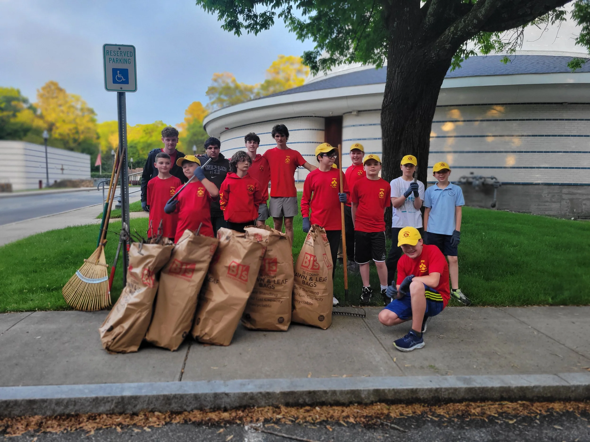 Group of Boy Scouts and young volunteers posing after a neighborhood cleanup, with trash bags, rakes, brooms, and gloves, in front of a building with a large tree.