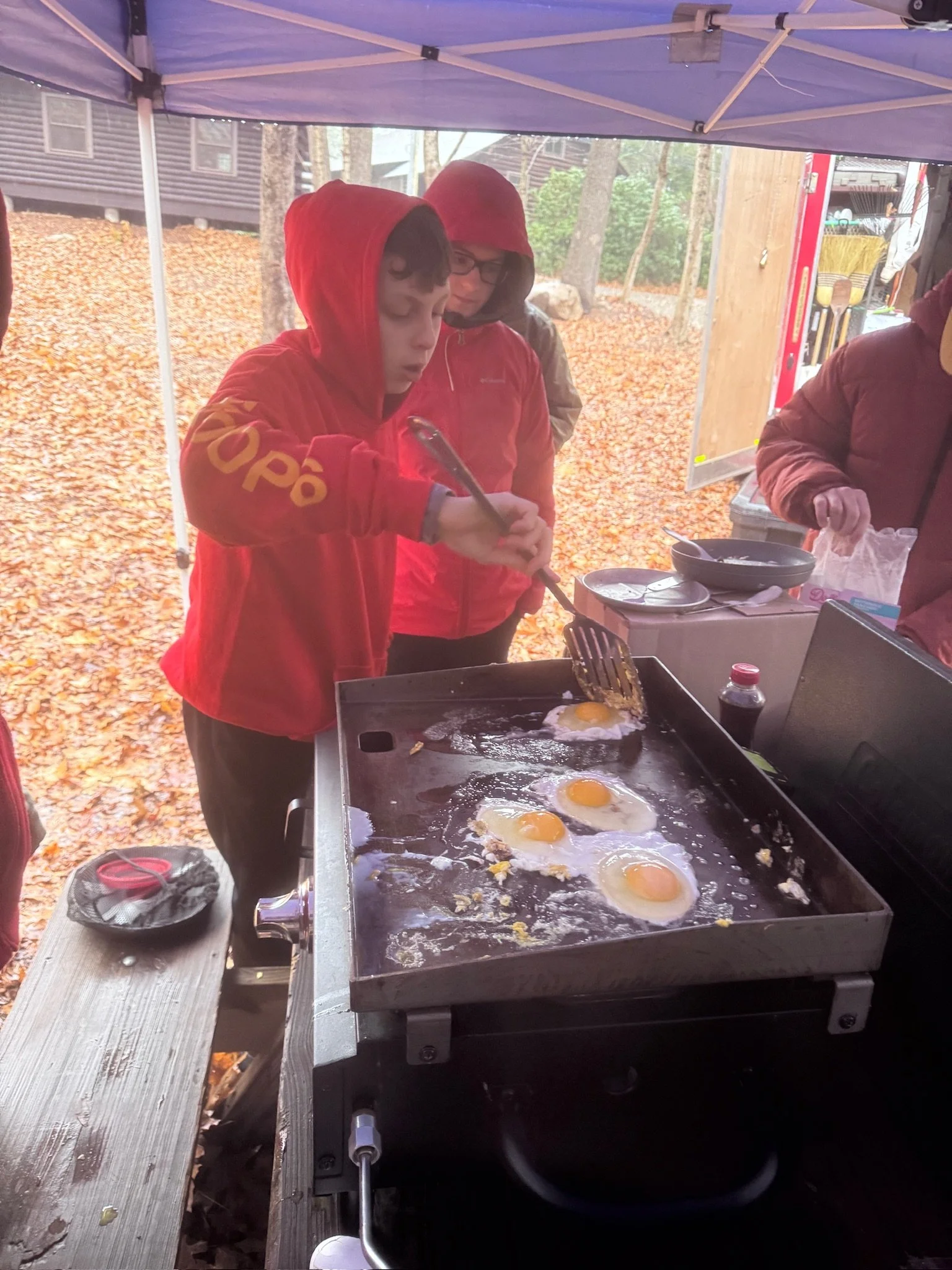 A boy wearing a red hoodie cooking sunny-side-up eggs on a griddle outdoors under a canopy, with two adults nearby in jackets and hoodies, surrounded by fallen autumn leaves.