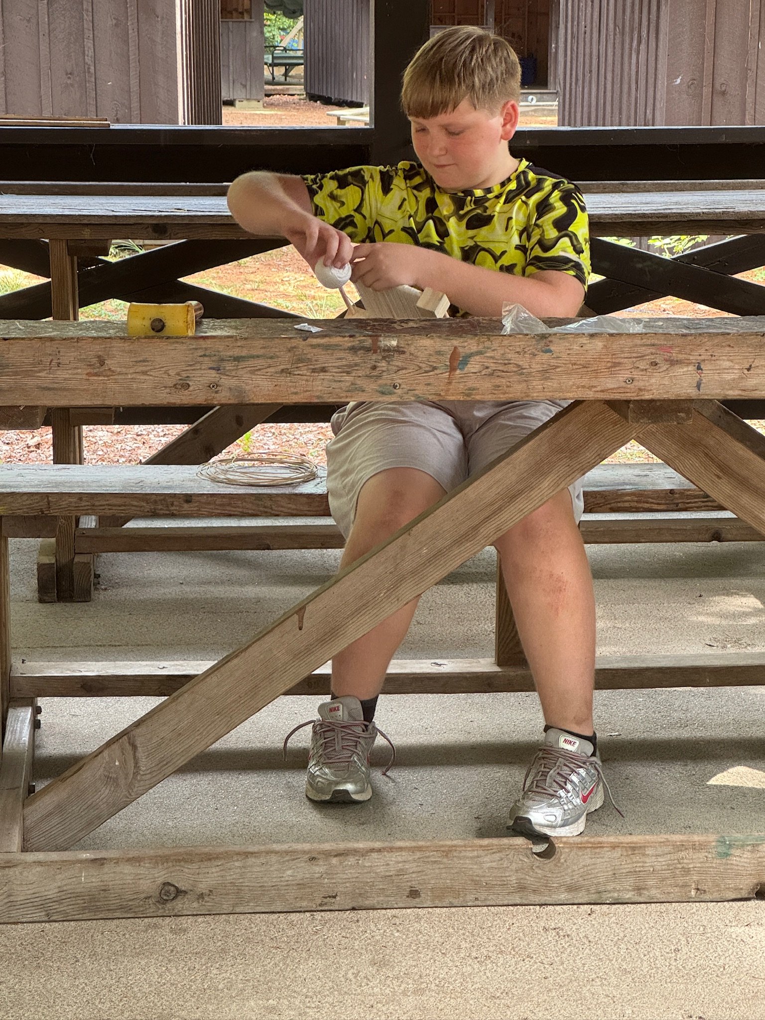 A boy sitting under a wooden picnic table, working on a craft project with glue and wooden pieces. The boy is wearing a yellow and black camouflage shirt, khaki shorts, and gray Nike sneakers.
