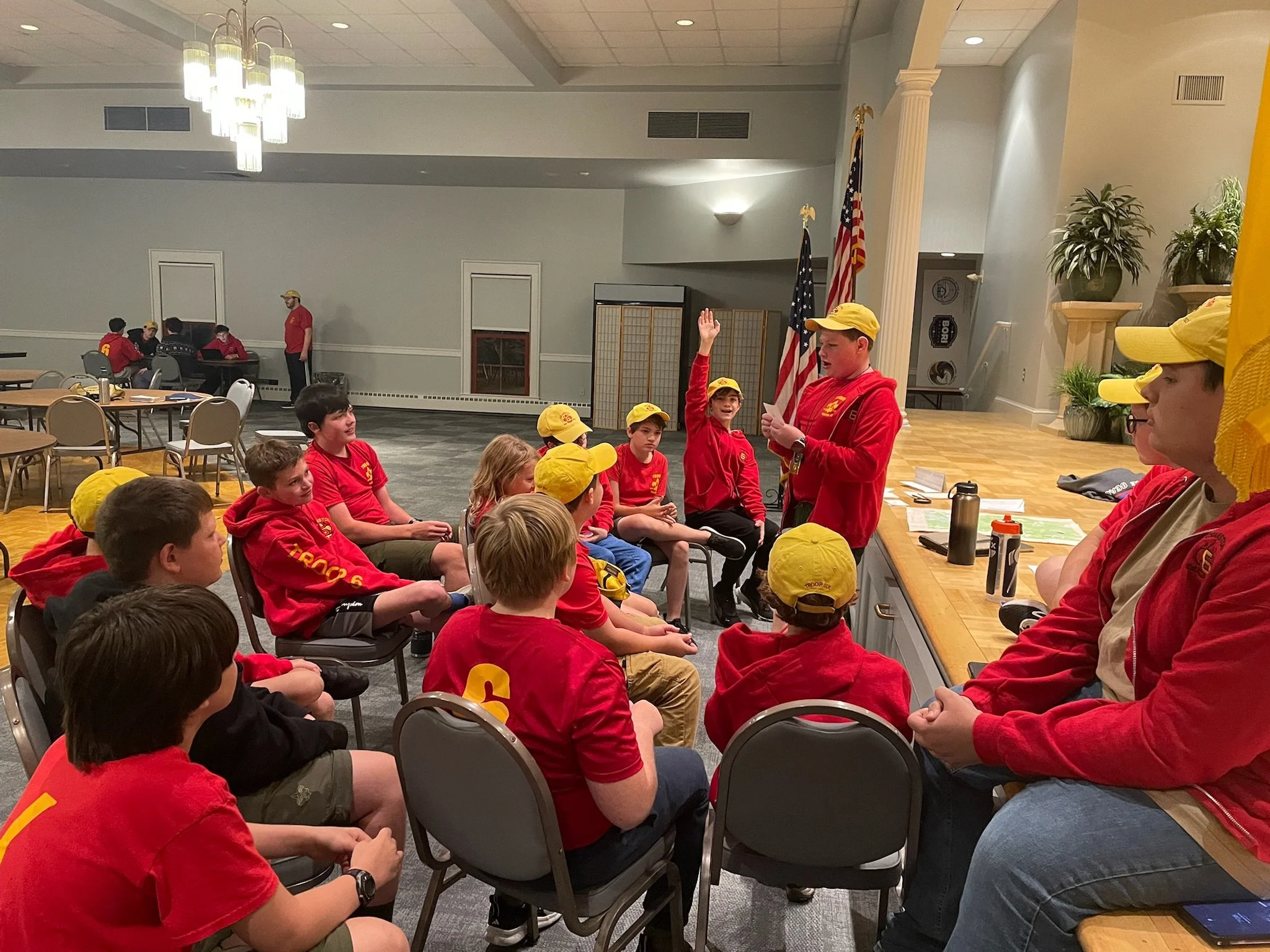 A group of children and teenagers in red and yellow uniforms attending a Boy Scouts meeting, seated and listening with a young boy raising his hand, with some adults in the background.  Meeting led by Scouts in our Scout Led program.