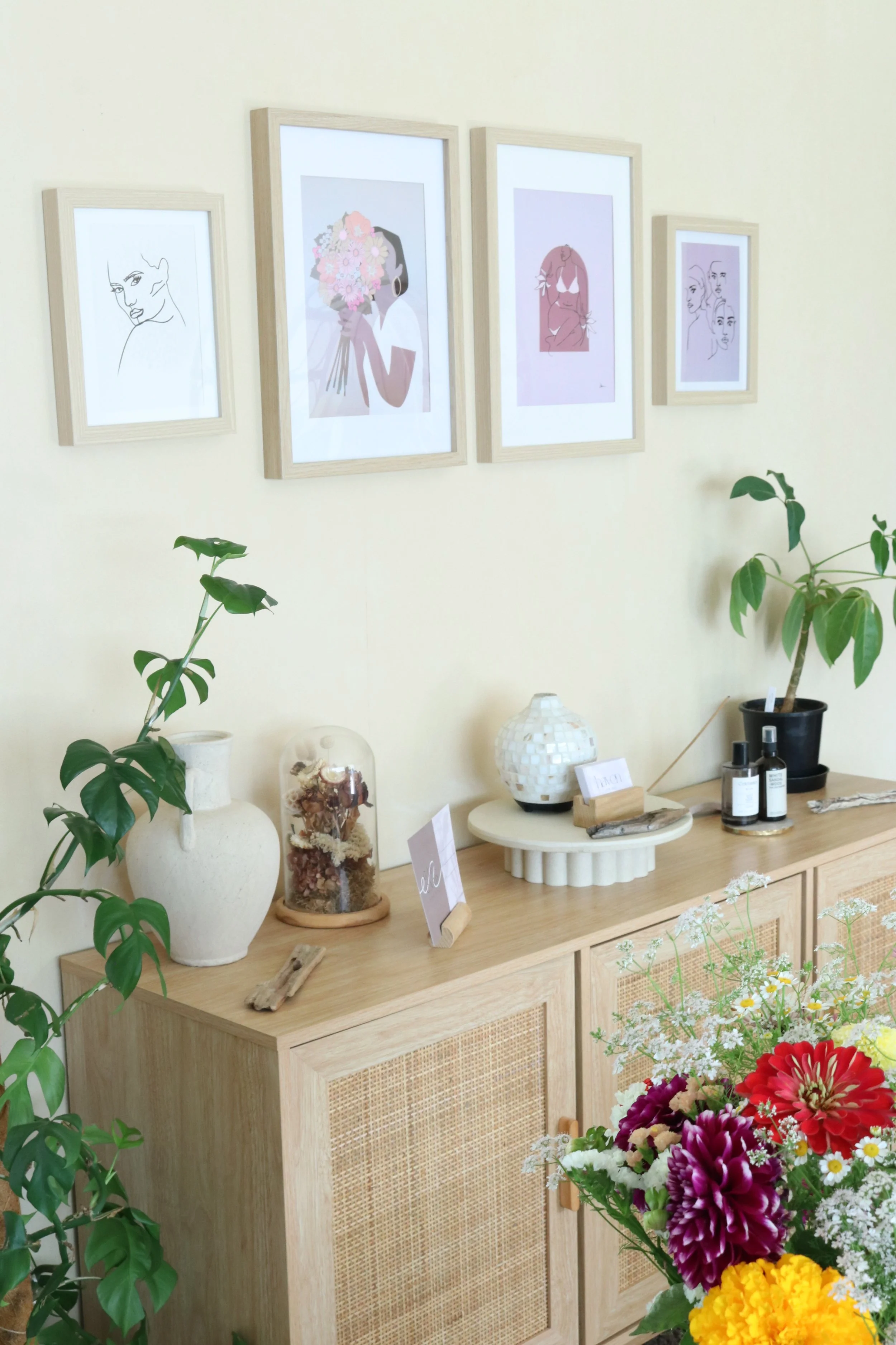 Interior of a room with framed artwork on the yellow wall, a wooden sideboard decorated with vases, plants, and decorative items, and a colorful bouquet of flowers in the foreground.