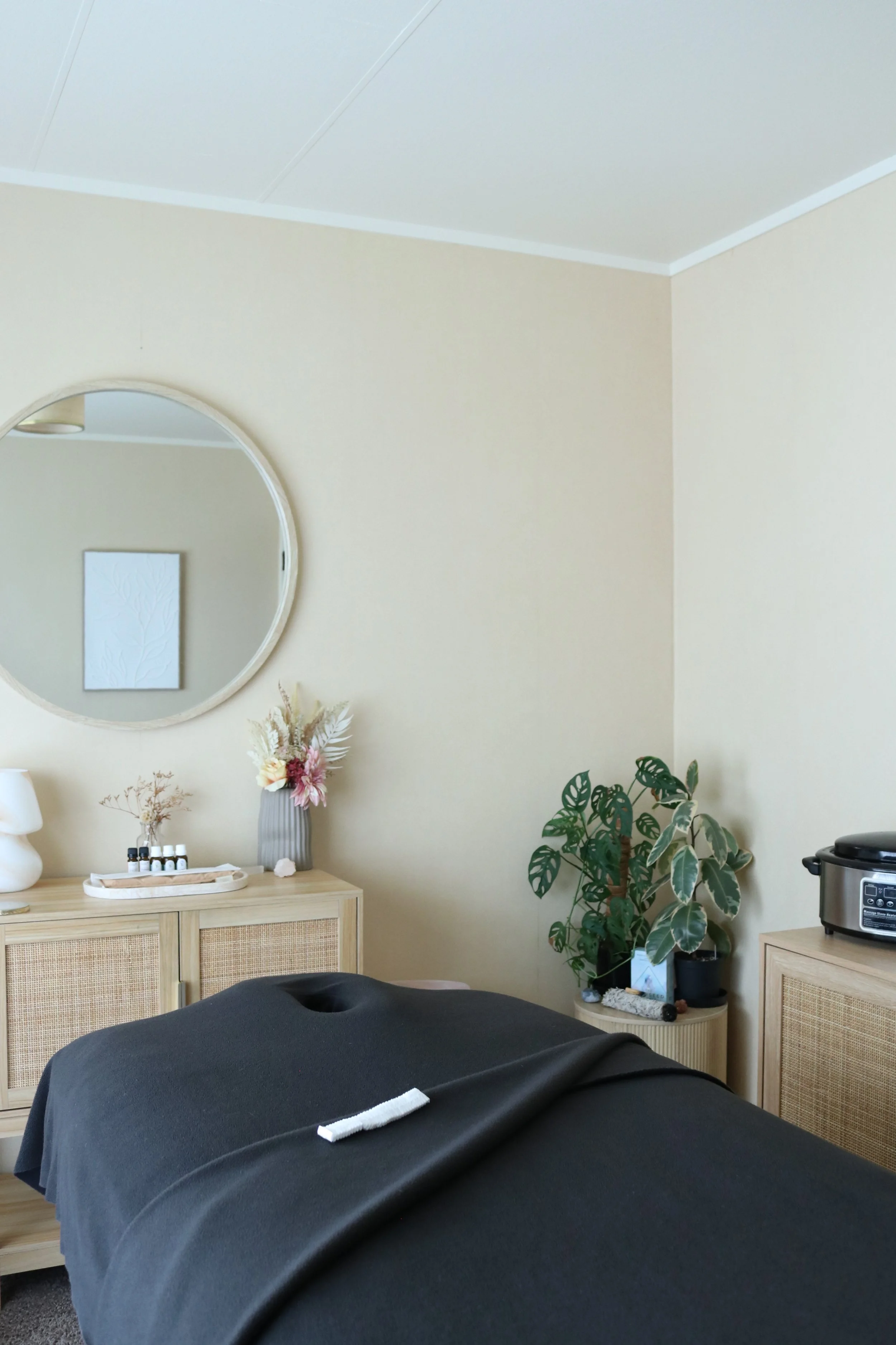 A cozy massage room with a massage table draped in a black sheet, a round mirror on the wall, a wooden sideboard with decorative items and essential oils, a potted plant, and a rice cooker on a cabinet.
