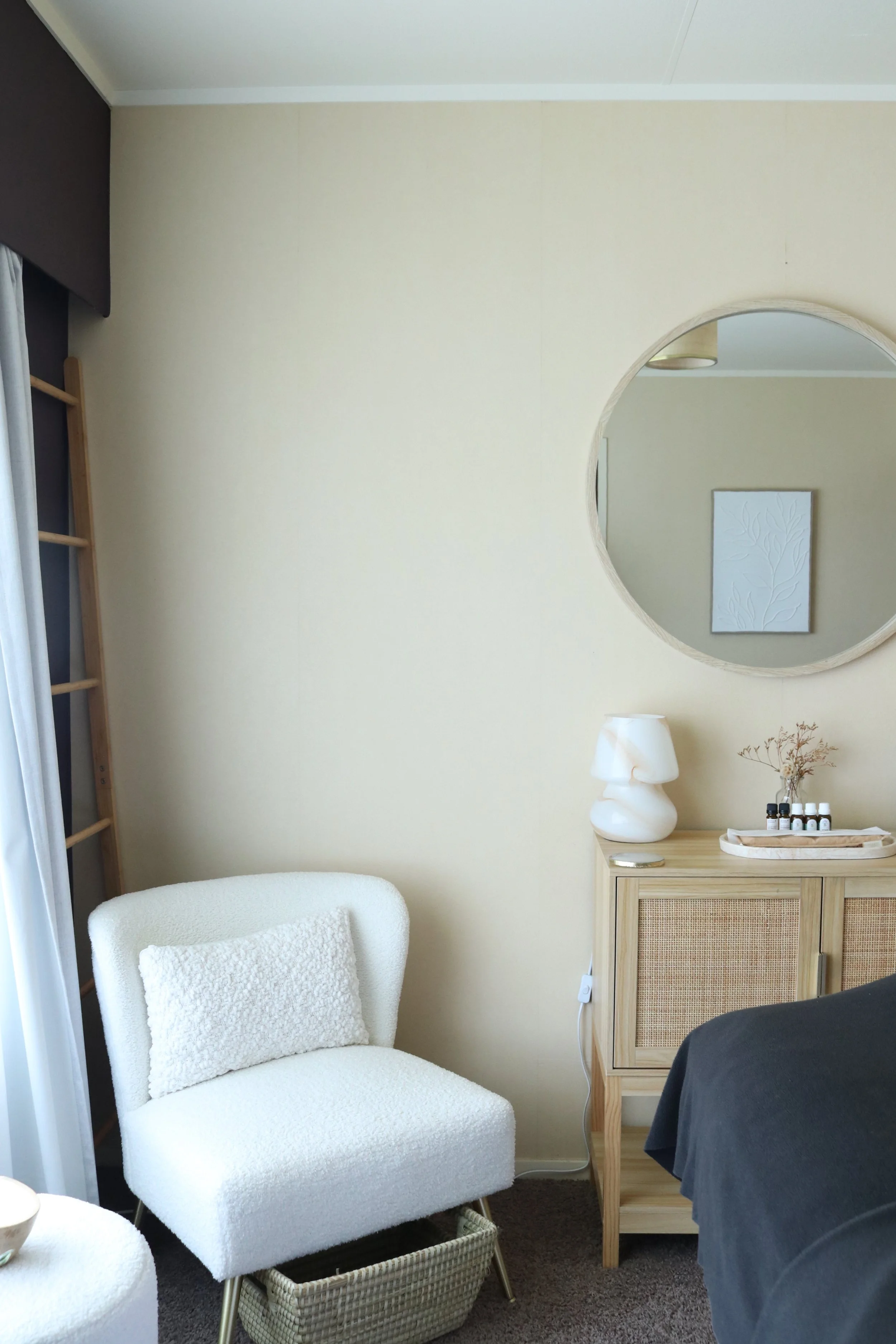 Interior of a room with a white upholstered chair, a wooden cabinet with decorative bottles and a vase, a large round mirror, and a dark bedspread.