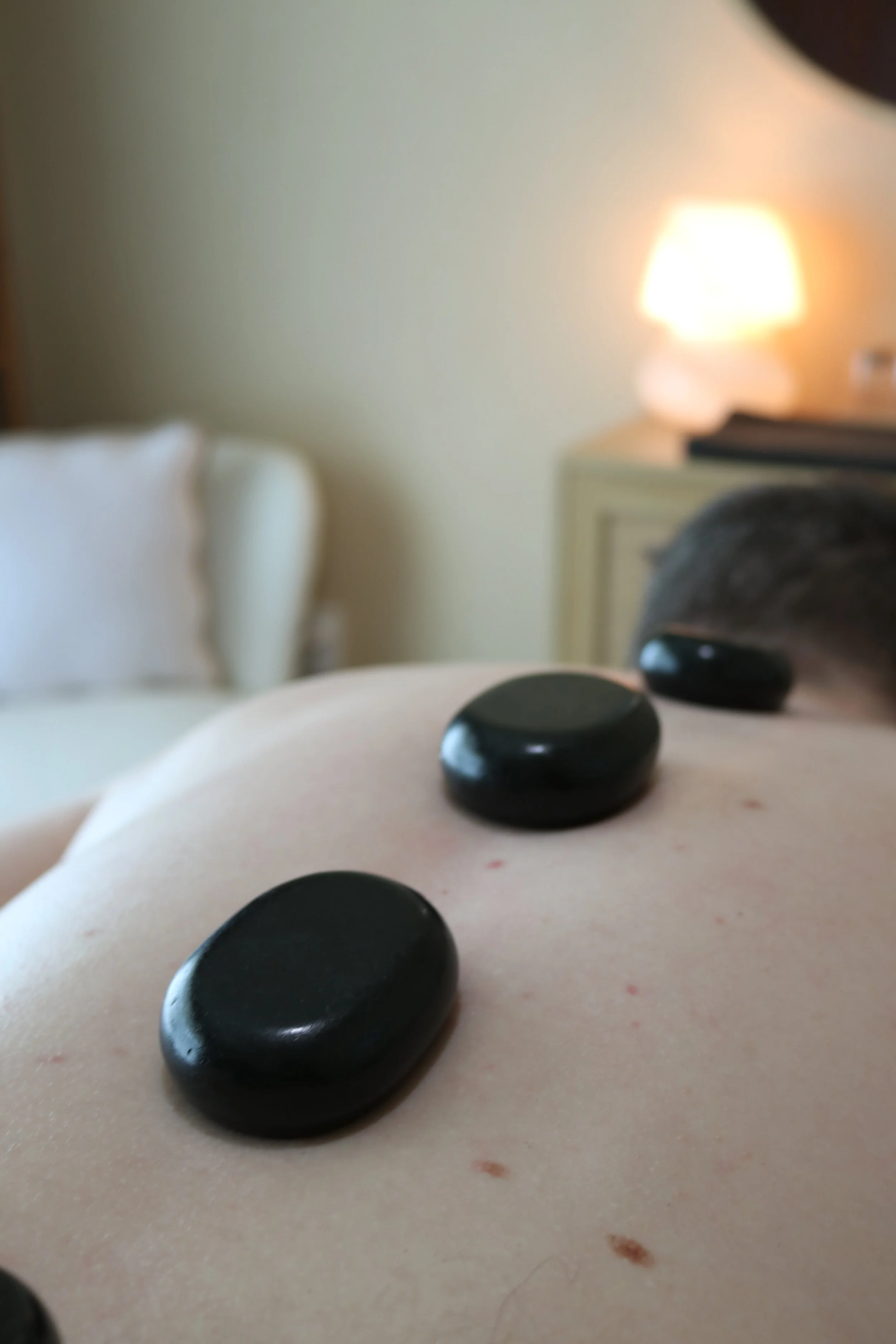 Close-up of hot stones placed on a person's back during a hot stone massage, with a blurred background of a bedside lamp and furniture in a tranquil room.