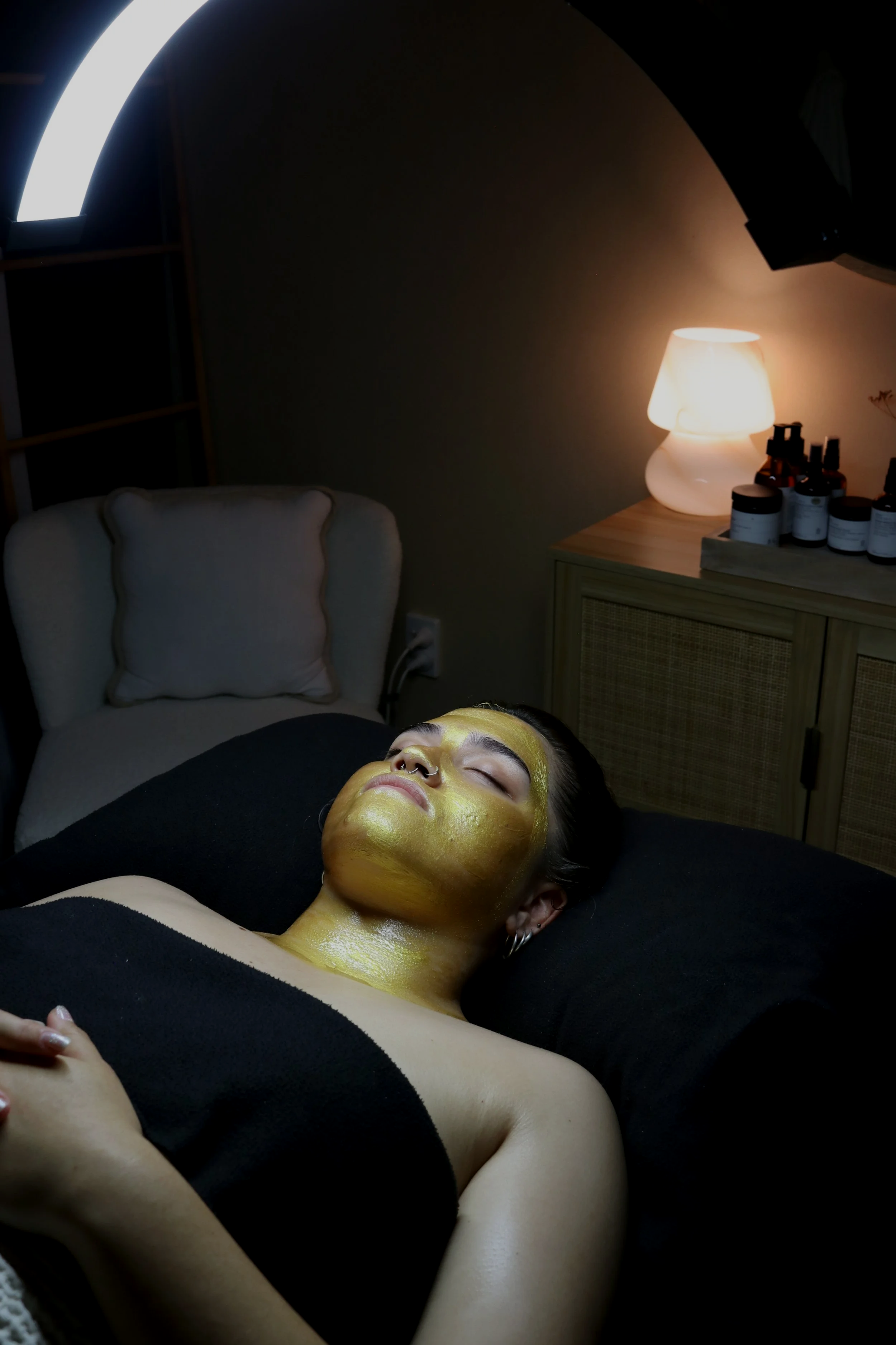 A woman lying down with a gold facial mask, relaxing with her eyes closed in a dimly lit spa or treatment room.