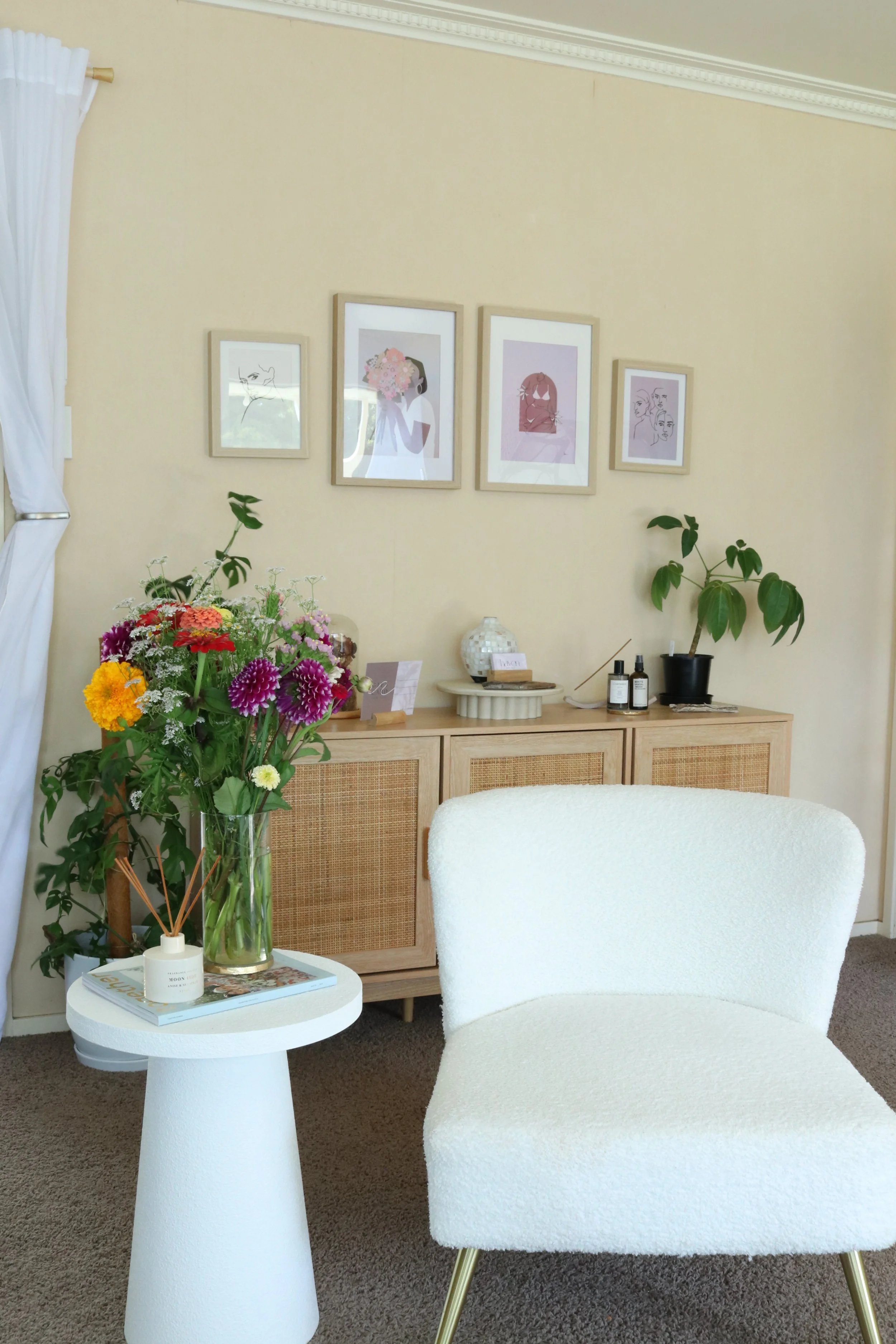 Interior of a room with a white chair, a white side table with a vase of colorful flowers, a framed wall art above a wooden cabinet, and a houseplant on the cabinet.