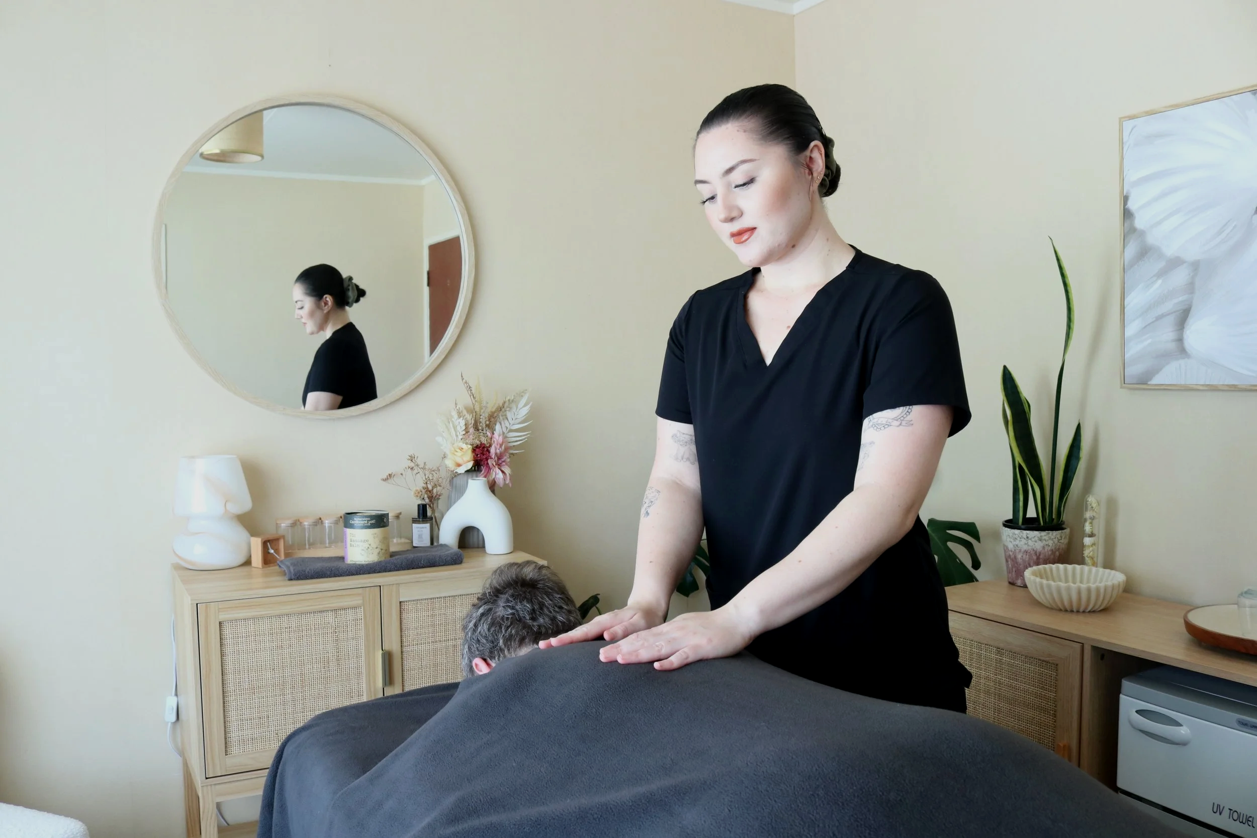 A woman in black scrubs giving a massage to a man lying face down on a massage table in a cozy room with beige walls. There is a large round mirror on the wall, a dresser with decorative items, plants, and artwork.