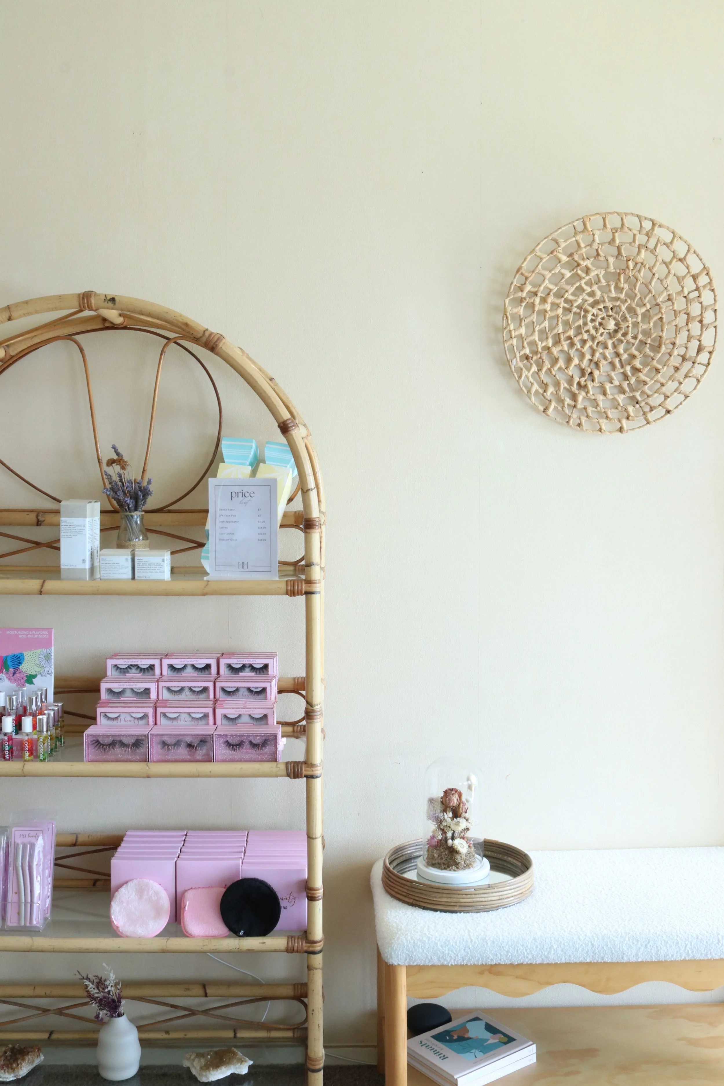 Interior of a store with rattan shelving holding cosmetics, lashes, and skincare products. A small bench with a decorative tray and a book underneath, along with wall decor including a circular woven piece.