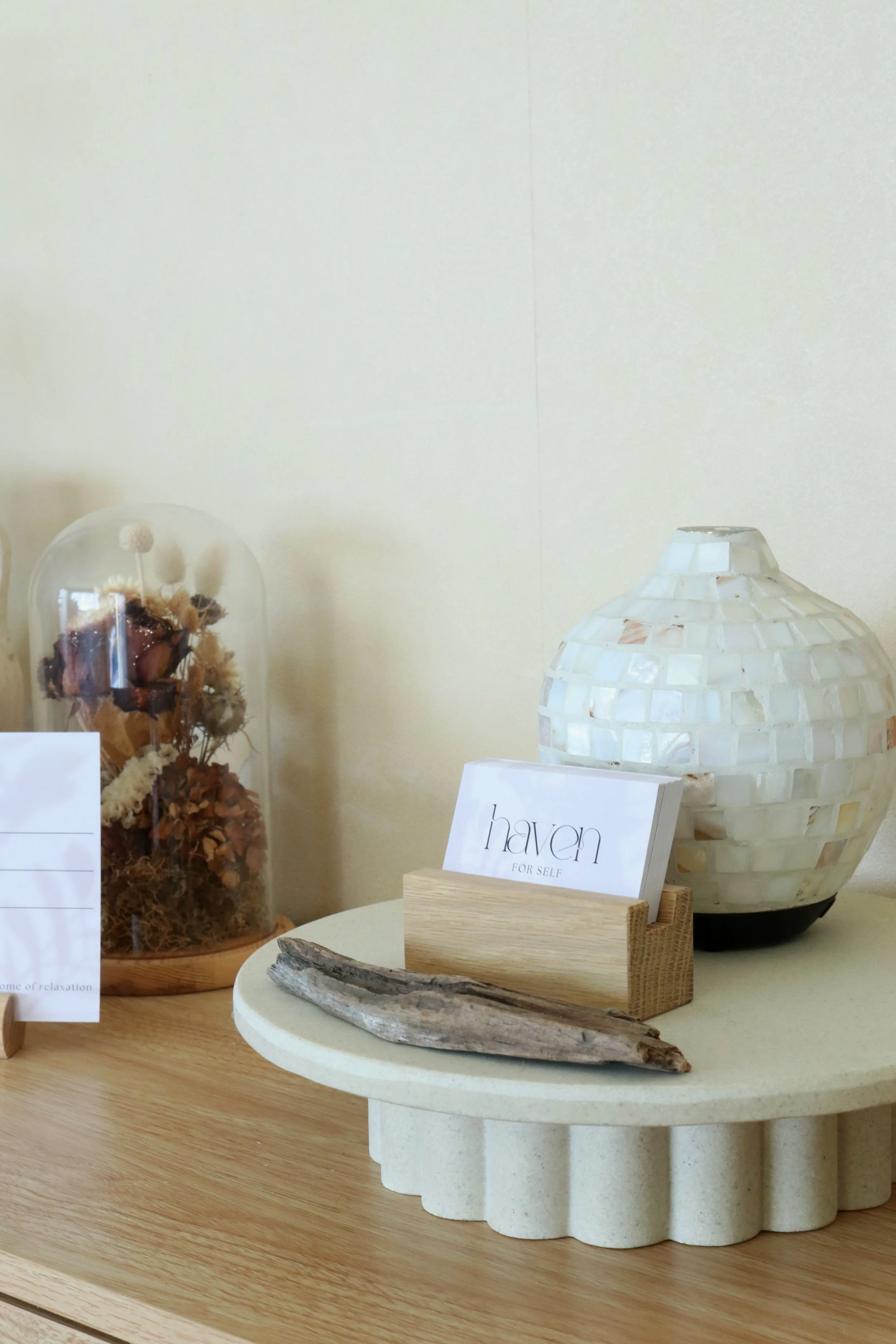 Decorative objects on a light-colored table, including a dried flower arrangement under a glass cover, a small cardholder with a card that reads "haven for self," a long piece of driftwood, and a round mosaic-style ceramic lamp, with a plain wall in the background.