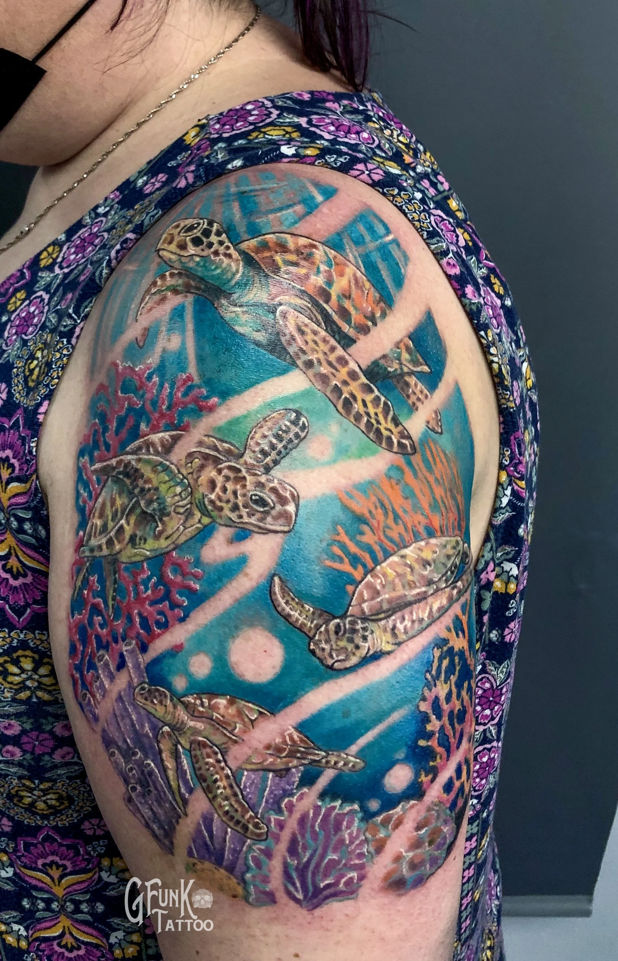 Colorful tattoo of sea turtles and coral on person's upper arm, with background of blue ocean and bubbles, wearing a floral shirt and necklace.
