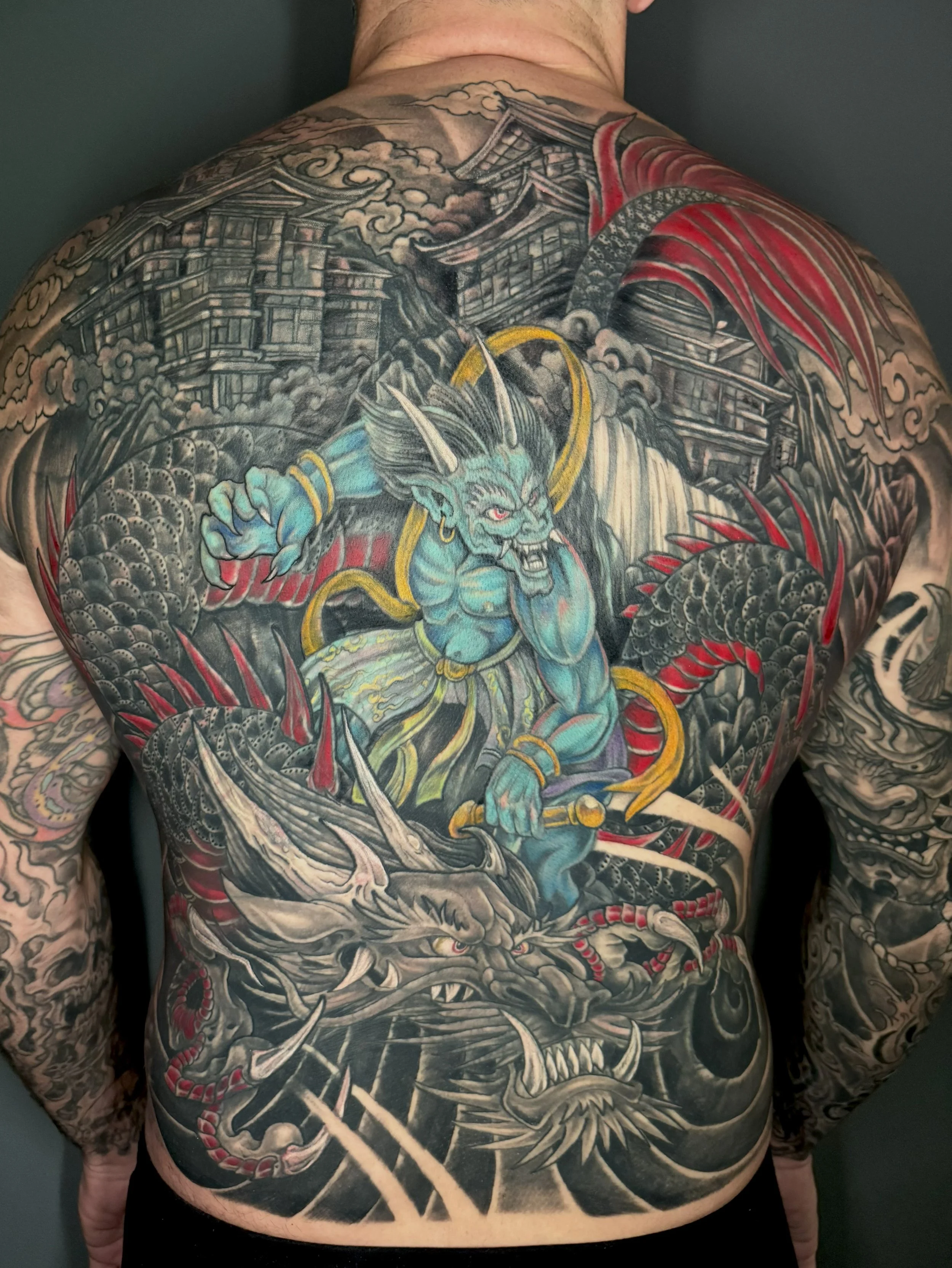 Tattoo of a fierce warrior with horns, wielding a weapon, surrounded by traditional Japanese architecture and swirling clouds, covering the person's back and arms.
