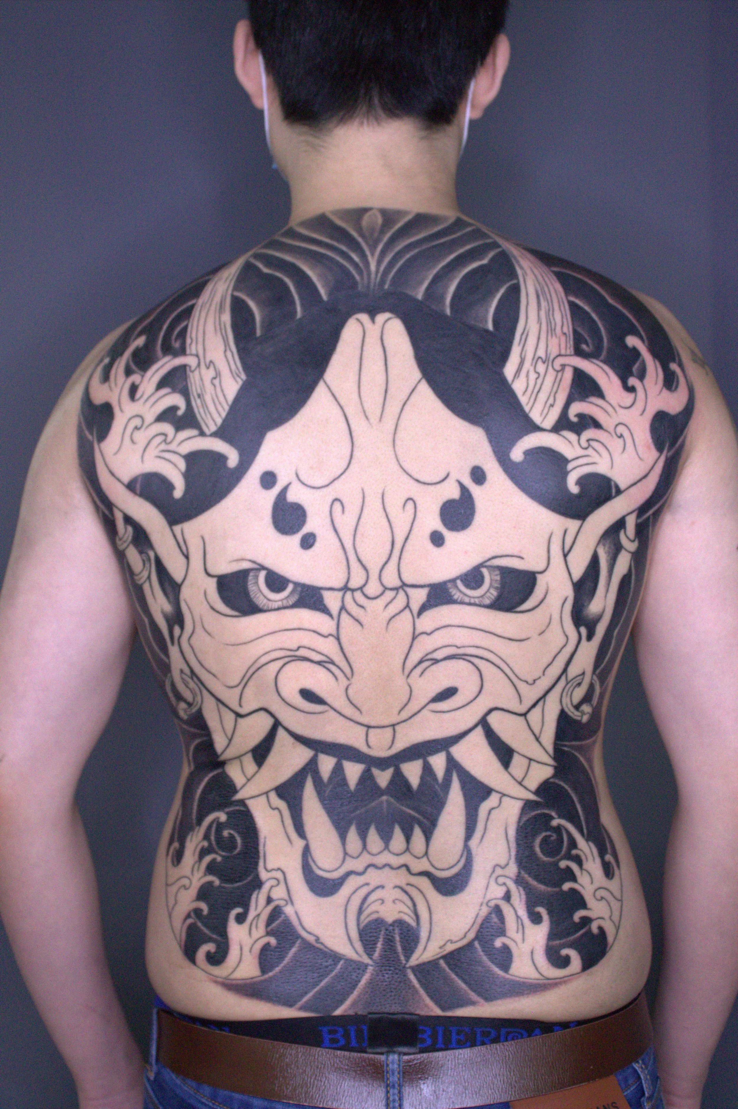 A person with a large black and beige tattoo of a fierce animal face, possibly a tiger or lion, covering the chest and shoulders. The person is shirtless, wearing jeans and a brown belt.