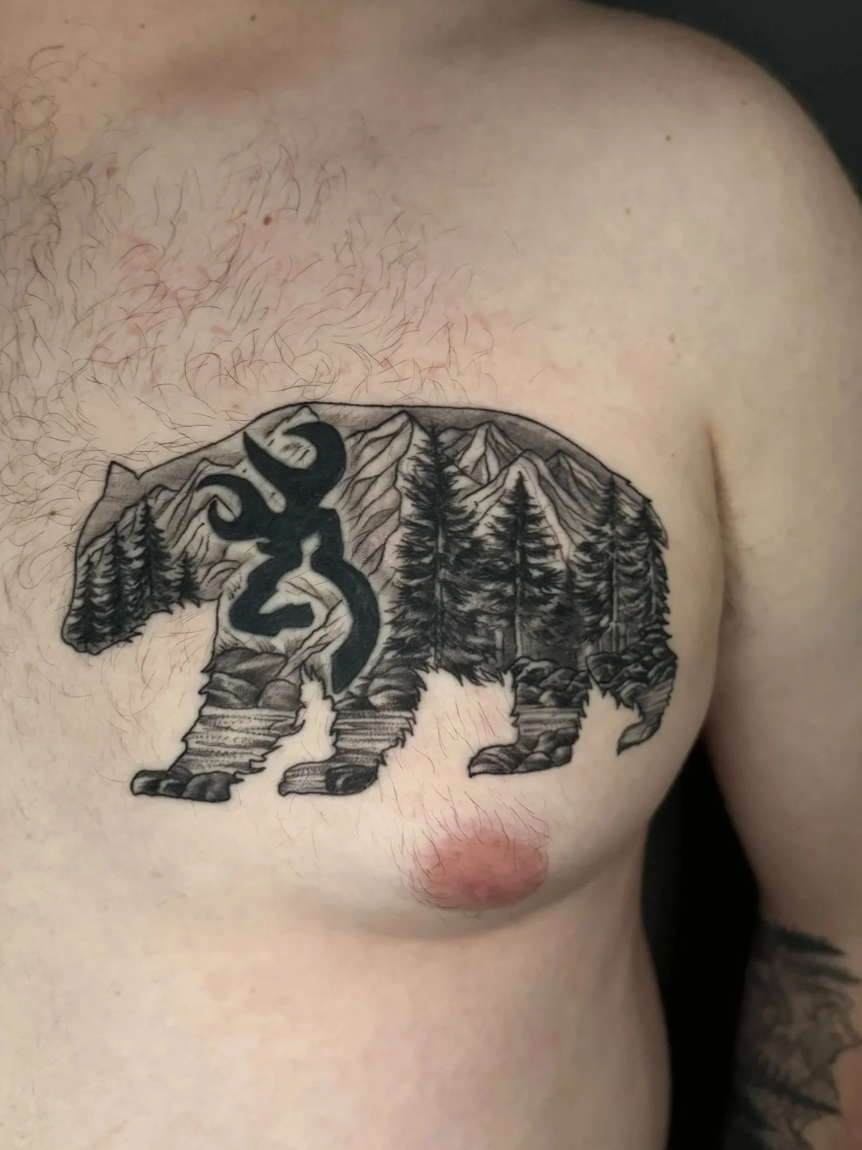 Black and gray tattoo of a bear with a forest scene, mountains, and trees on a person's chest.