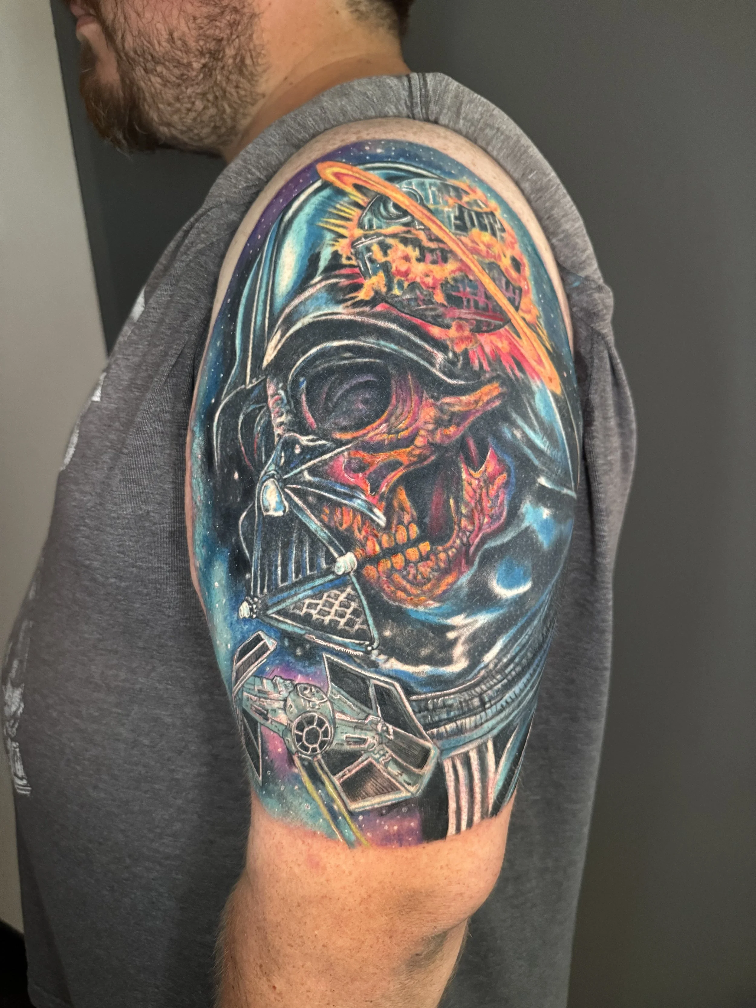 Colorful tattoo of a Darth Vader helmet with a skull inside, a spaceship, and space elements on a man's upper arm.