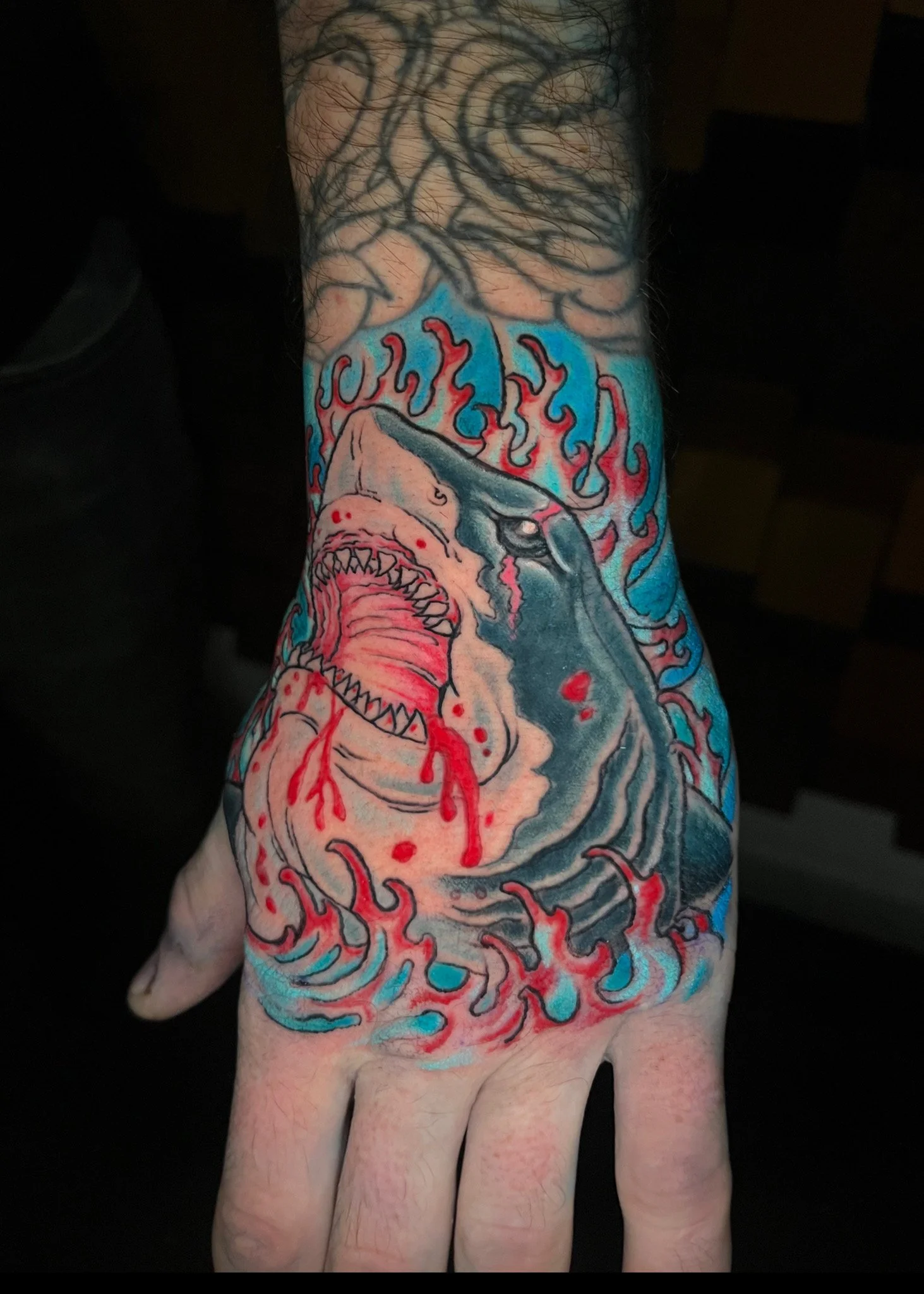 Tattoo of a shark with blood and flames on a person's hand and wrist.