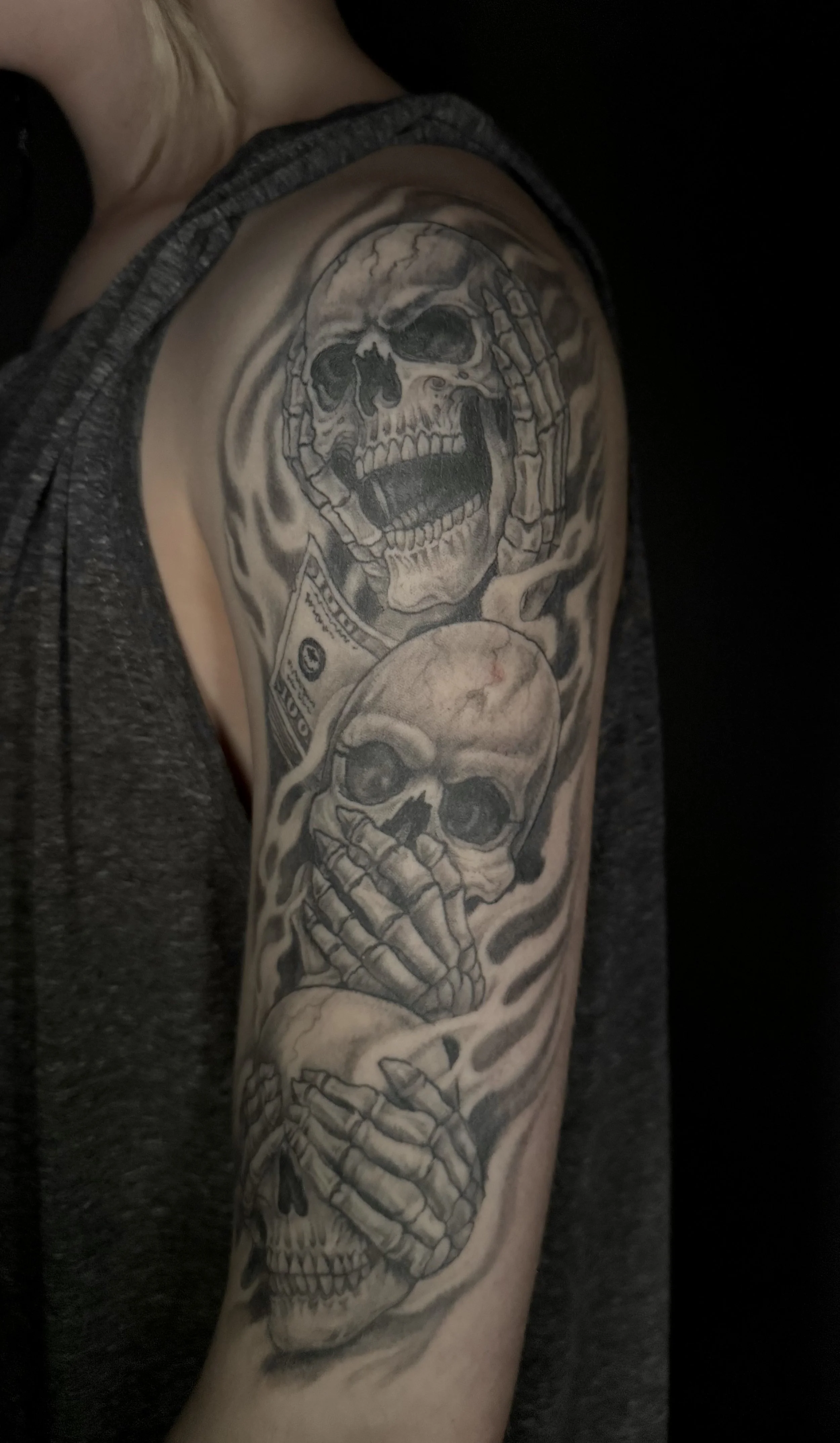 Tattoo of three skulls on person's upper arm, with skeleton hands covering their faces and a dollar bill in the background.